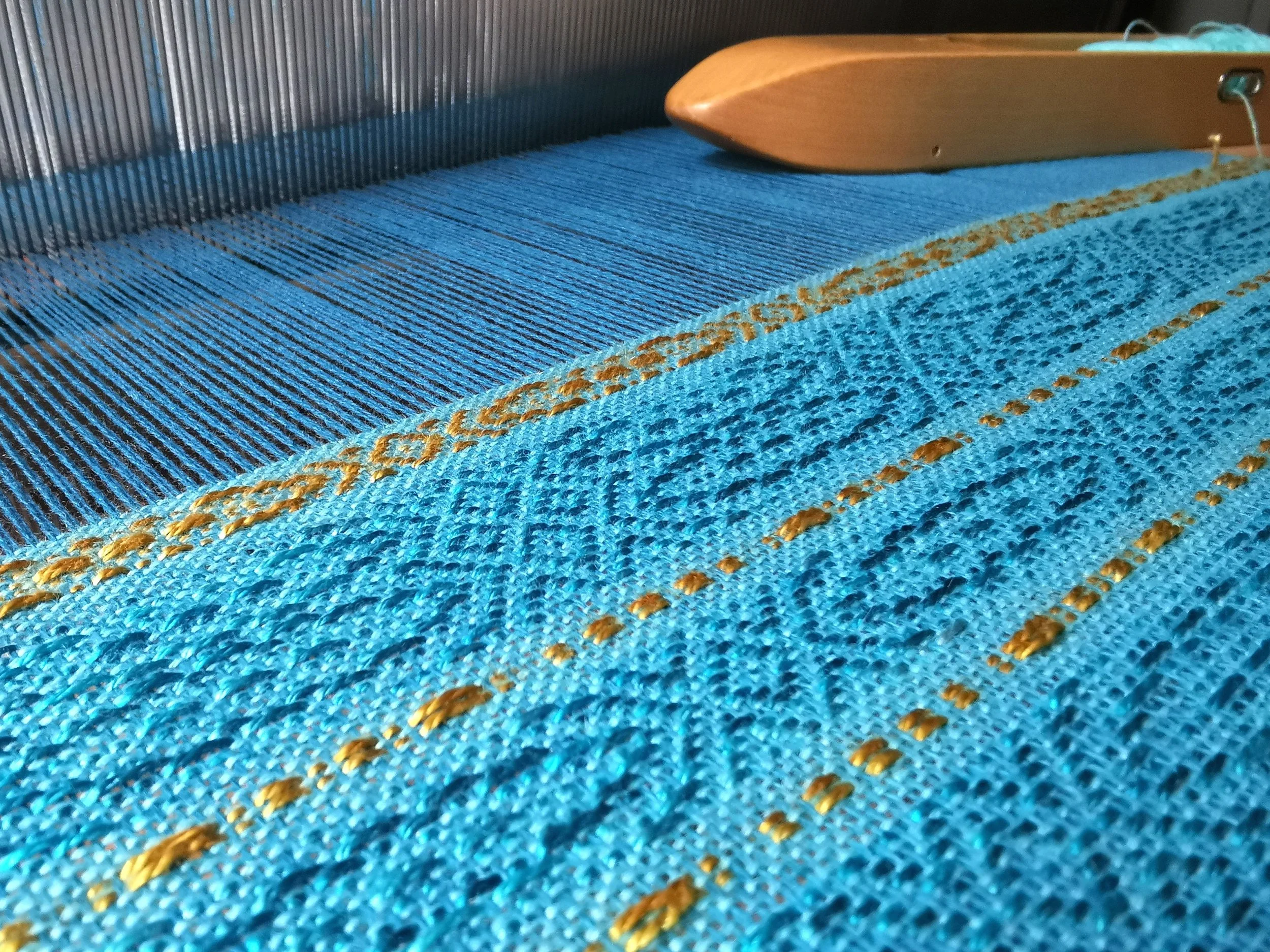 Gros plan sur l'étole Calypso en cours de tissage sur le métier, motif overshot turquoise et bandes de fil doré, fils de chaîne tendus