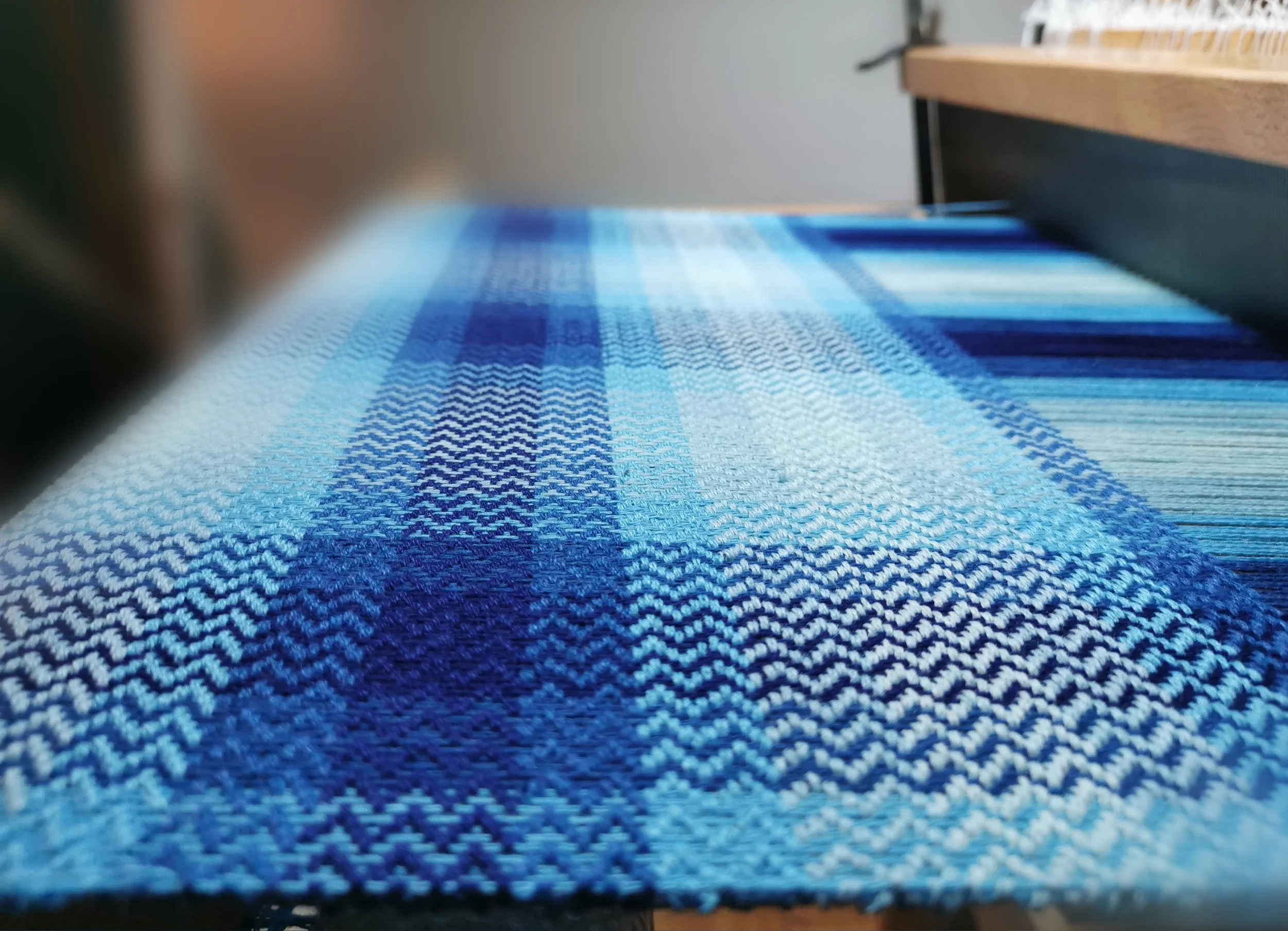 Gros plan sur l'écharpe Source en cours de tissage sur le métier, motif de carreaux en nuances de bleu et turquoise, fils de chaîne tendus