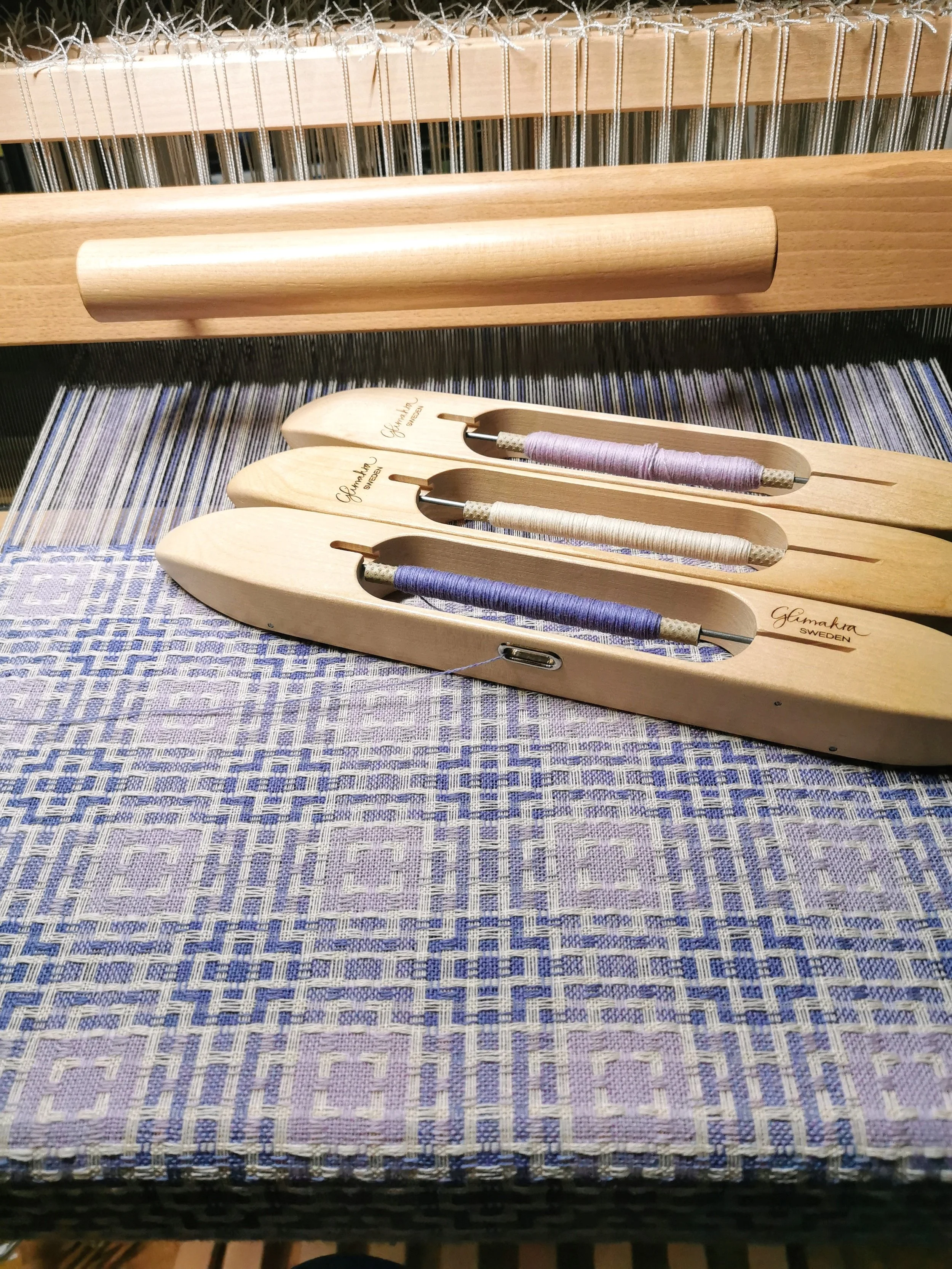 Écharpe Lilas en cours de tissage sur le métier : motif à petits carrés en violet foncé et violet pâle, trois navettes en bois garnies de fils violet clair, violet foncé et blanc