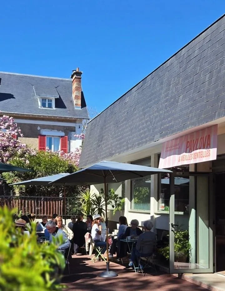 Groupe de personnes assises à une terrasse de café ou restaurant en plein air, avec un parasol, près d'une bâtisse moderne, sous un ciel bleu clair.