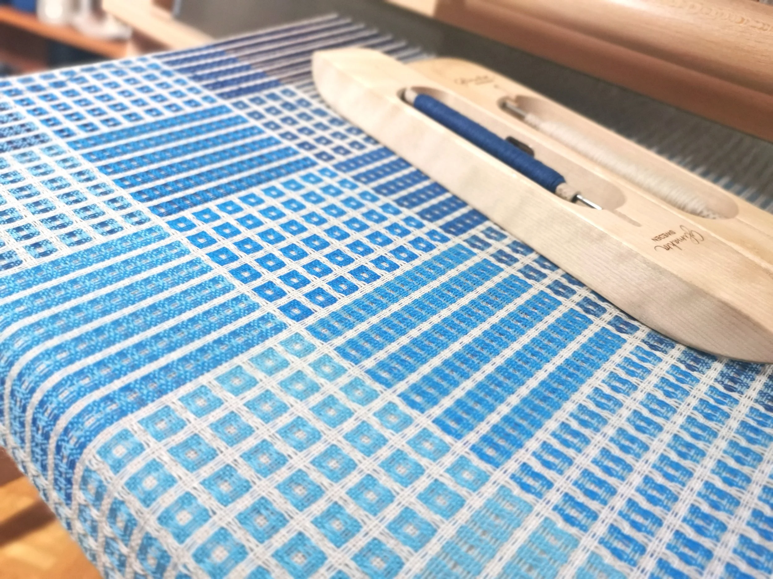 Écharpe Variation en bleu en cours de tissage sur le métier : zones de rayures et de petits motifs dans des bleus variés sur fond écru, navette en bois posée sur l'ouvrage