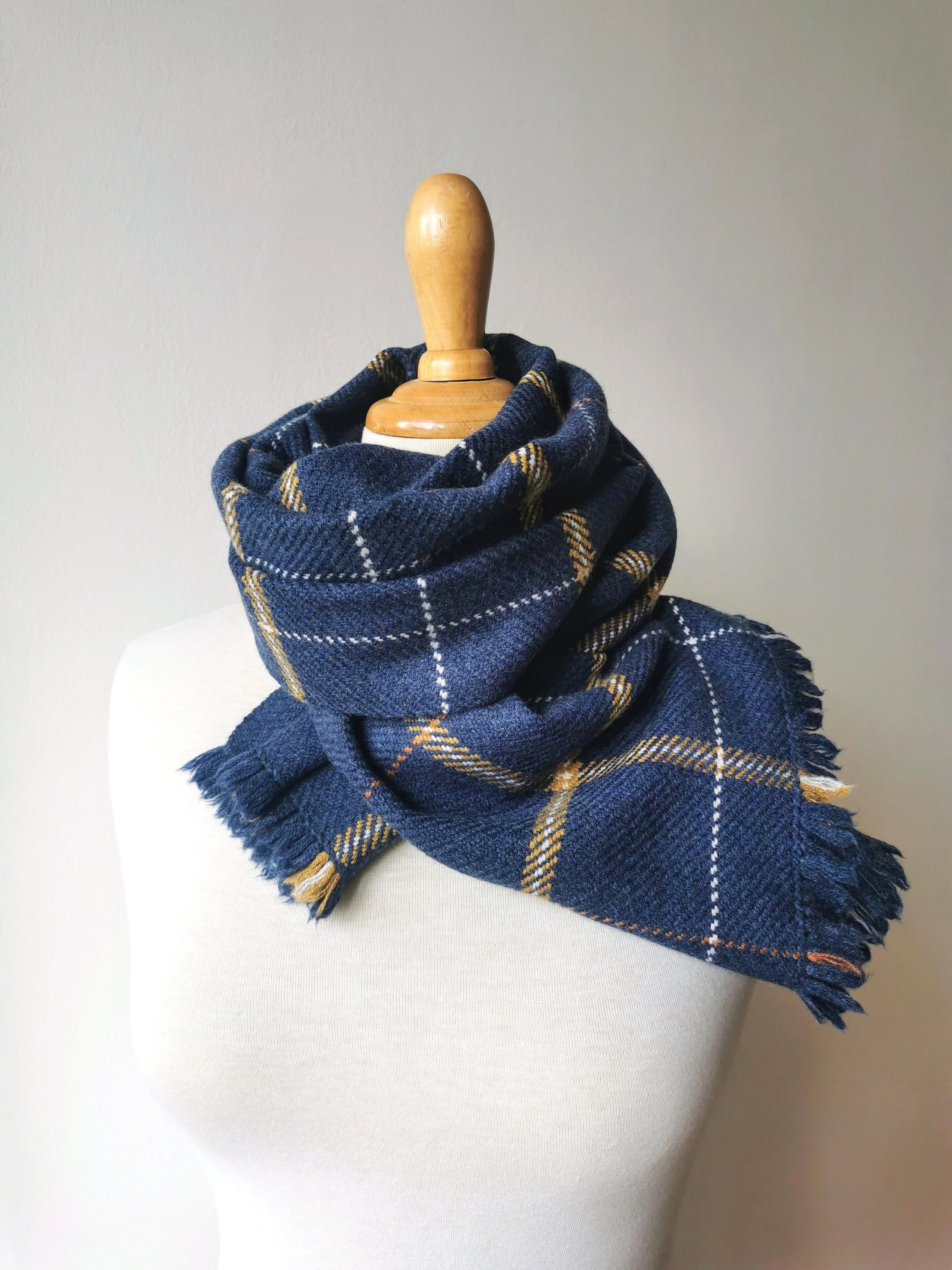 Écharpe Loch en laine mérinos roulée et nouée autour du cou d'un mannequin, motif de carreaux bleu profond avec lignes écrues, orangées et dorées, franges visibles