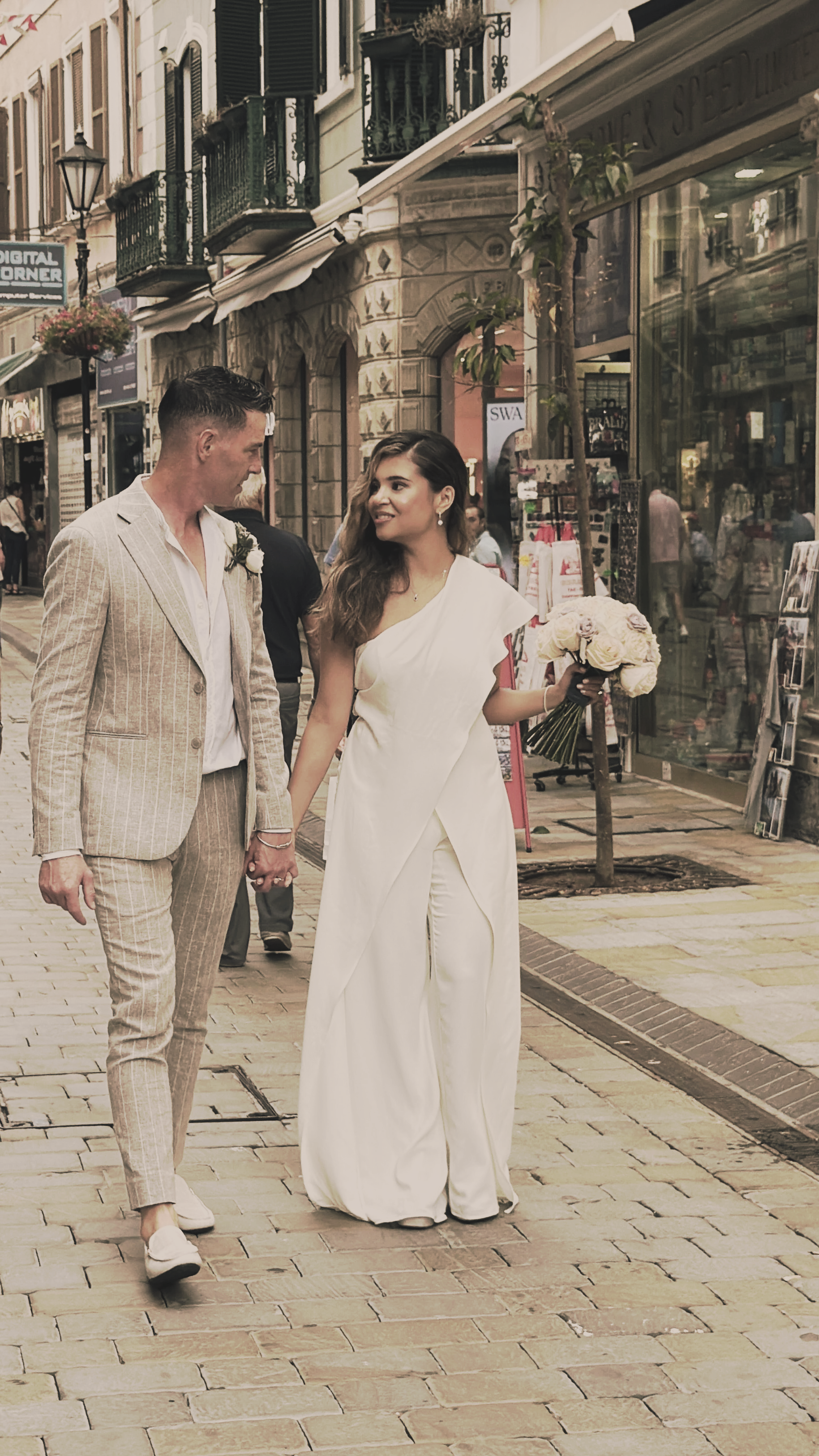 A couple dressed in wedding attire walking hand-in-hand on a city street, with the woman holding a bouquet of white roses.
