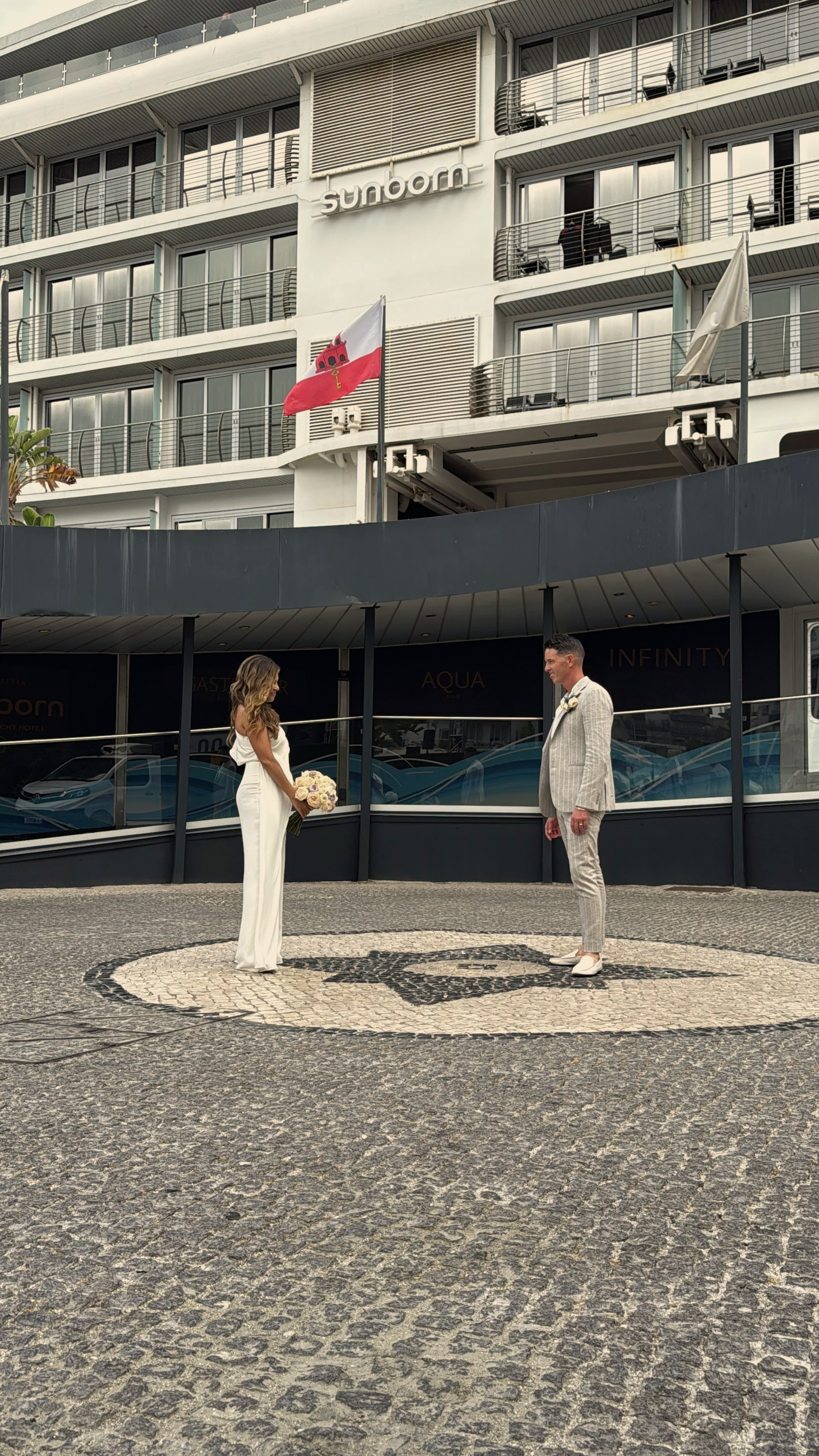 A bride and groom standing face-to-face outdoors in front of a hotel, with the bride holding a bouquet of flowers. The hotel building behind them has the name 'Sunborn' and flags, one of which is red and white with an emblem. The scene appears to be part of a wedding ceremony.