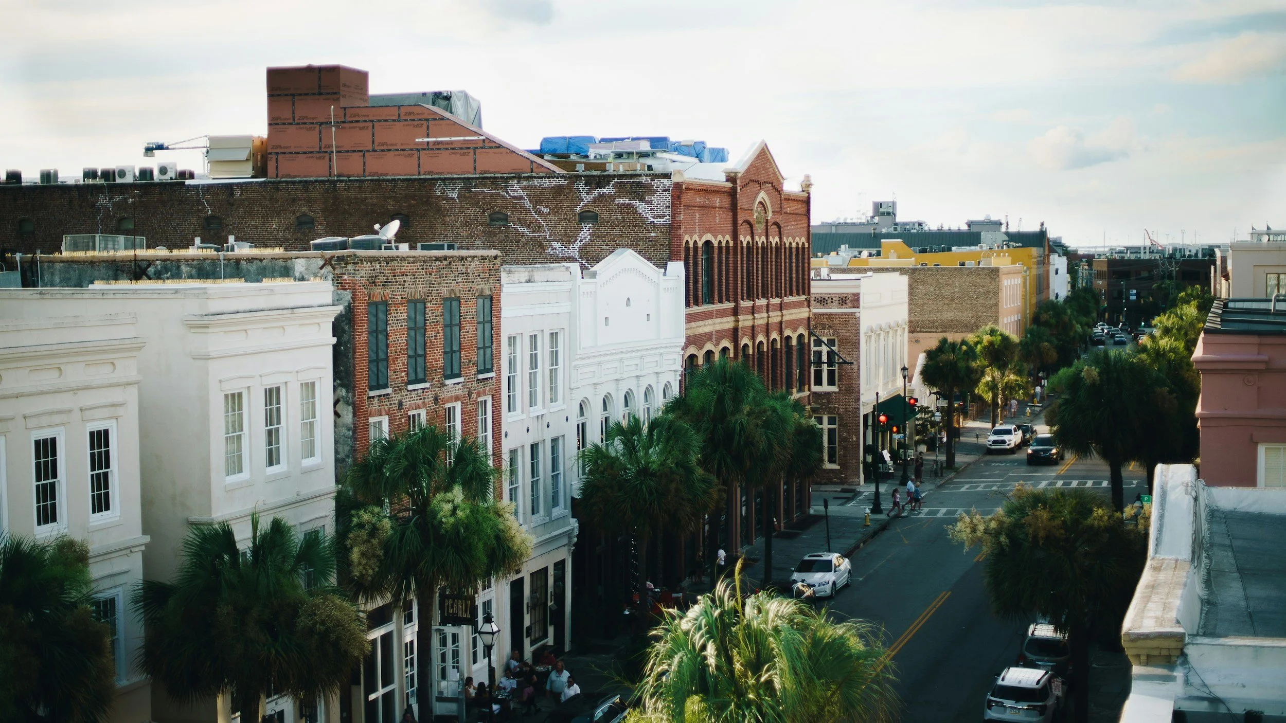 View of a city street lined with historic buildings, palm trees, and parked cars, with pedestrians walking along the sidewalk under a partly cloudy sky.