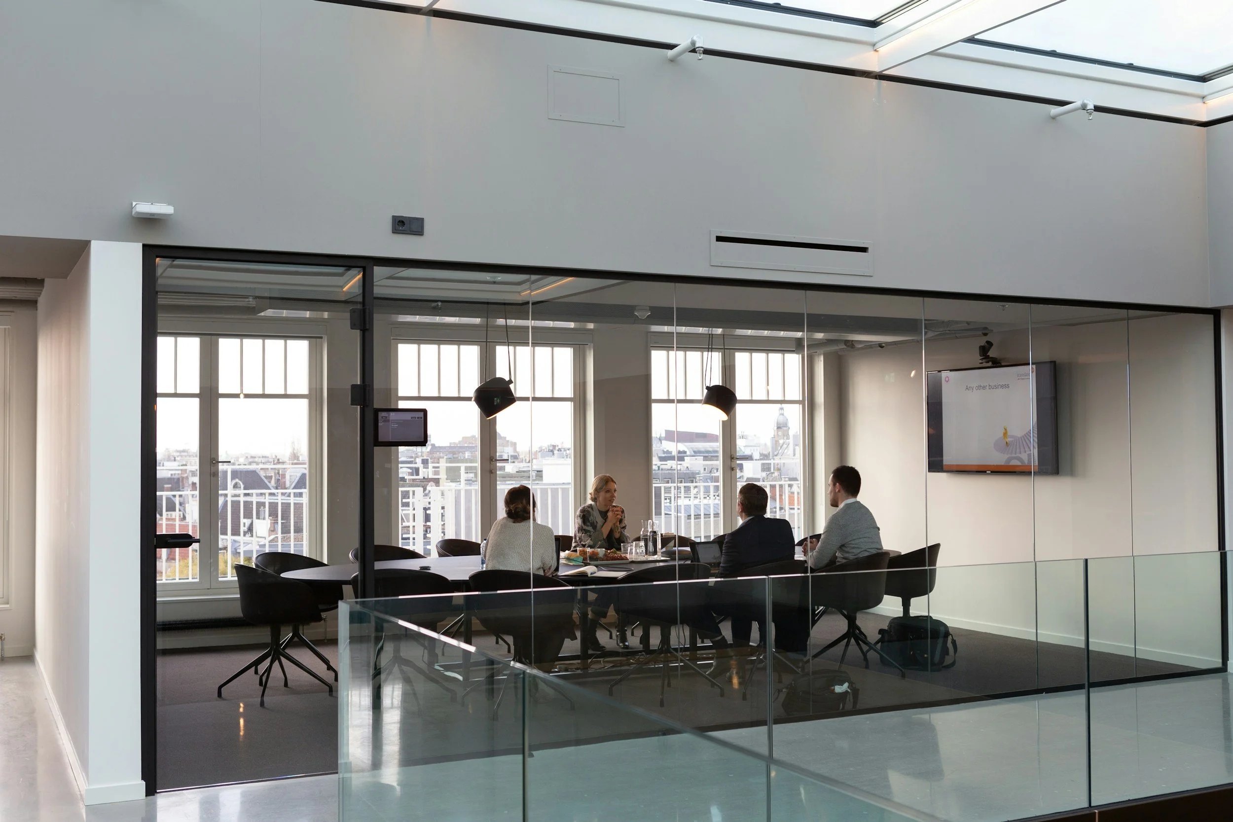 A modern conference room with five people seated around a table, having a discussion. The room has large windows with city views and a flat screen TV on the wall displaying a presentation.