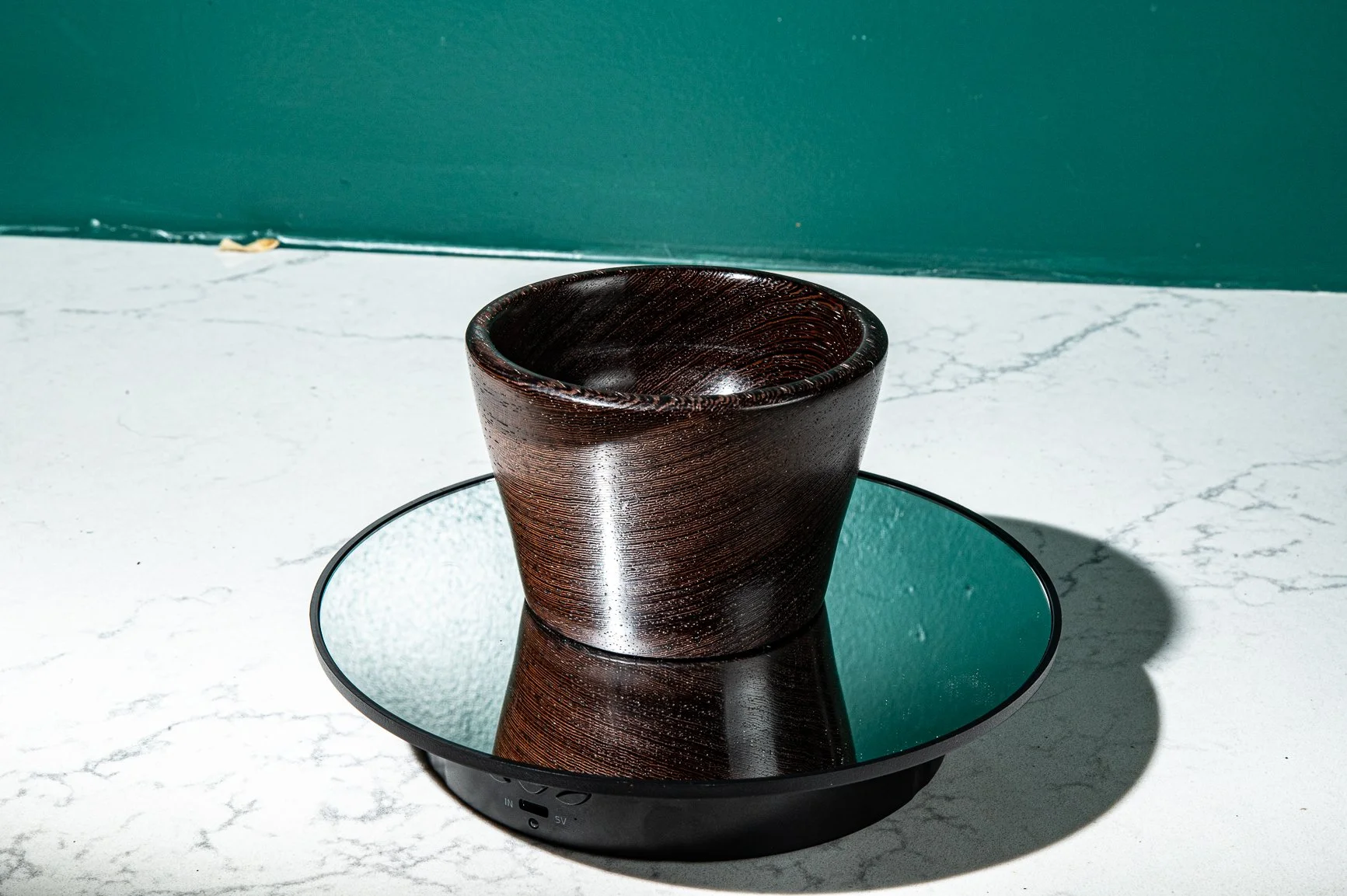 A dark wooden bowl placed on a round mirror tray on a white marble surface with a teal-colored wall in the background.