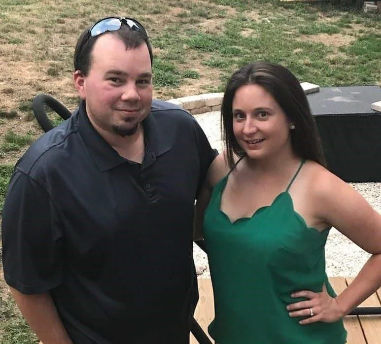 A man and woman standing outdoors, smiling and posing for the photo. The man wears a black shirt and sunglasses on his head. The woman wears a green top with spaghetti straps, has long dark hair, and a ring on her left hand.