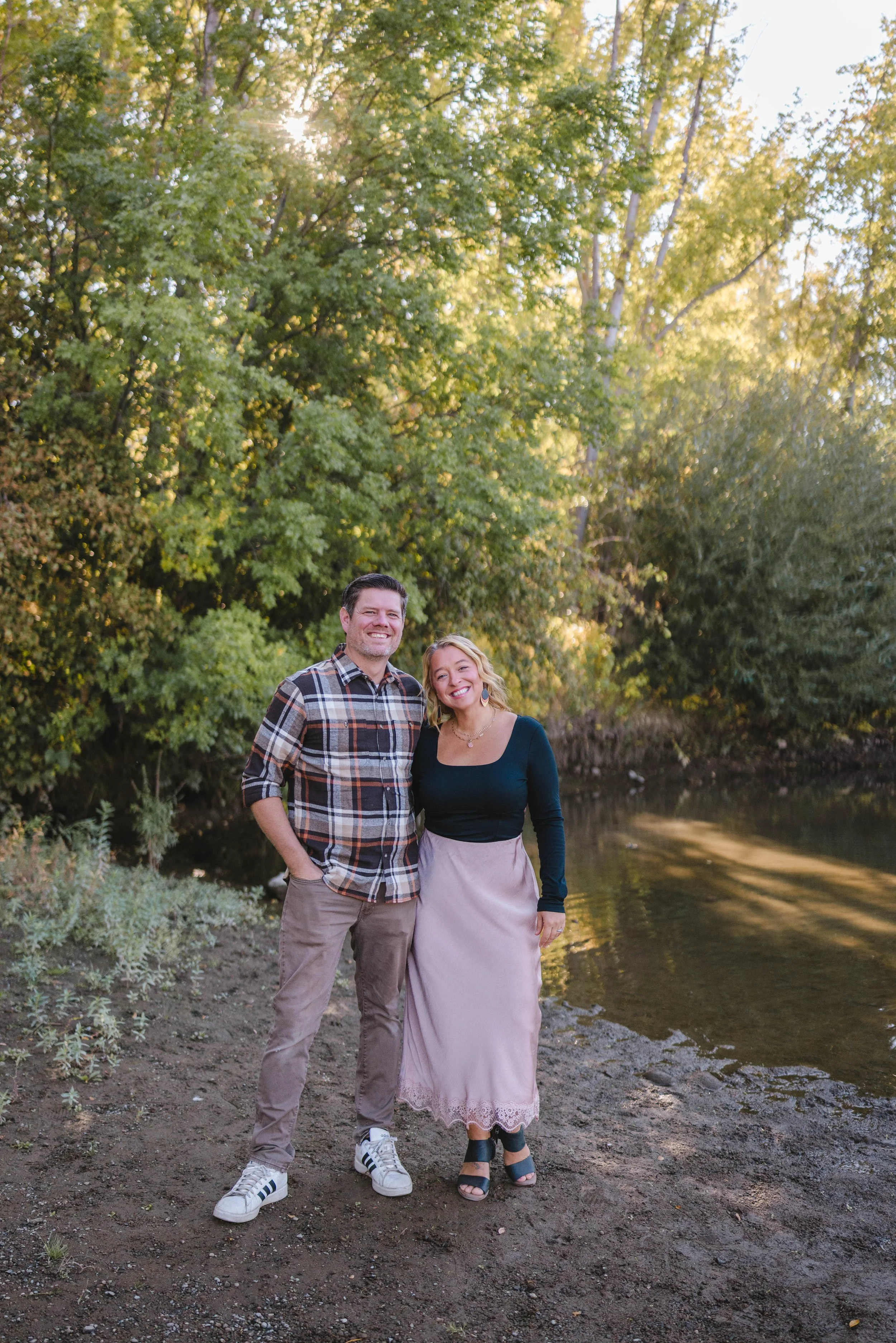 A smiling man and woman standing together near a river in a wooded area during sunset.