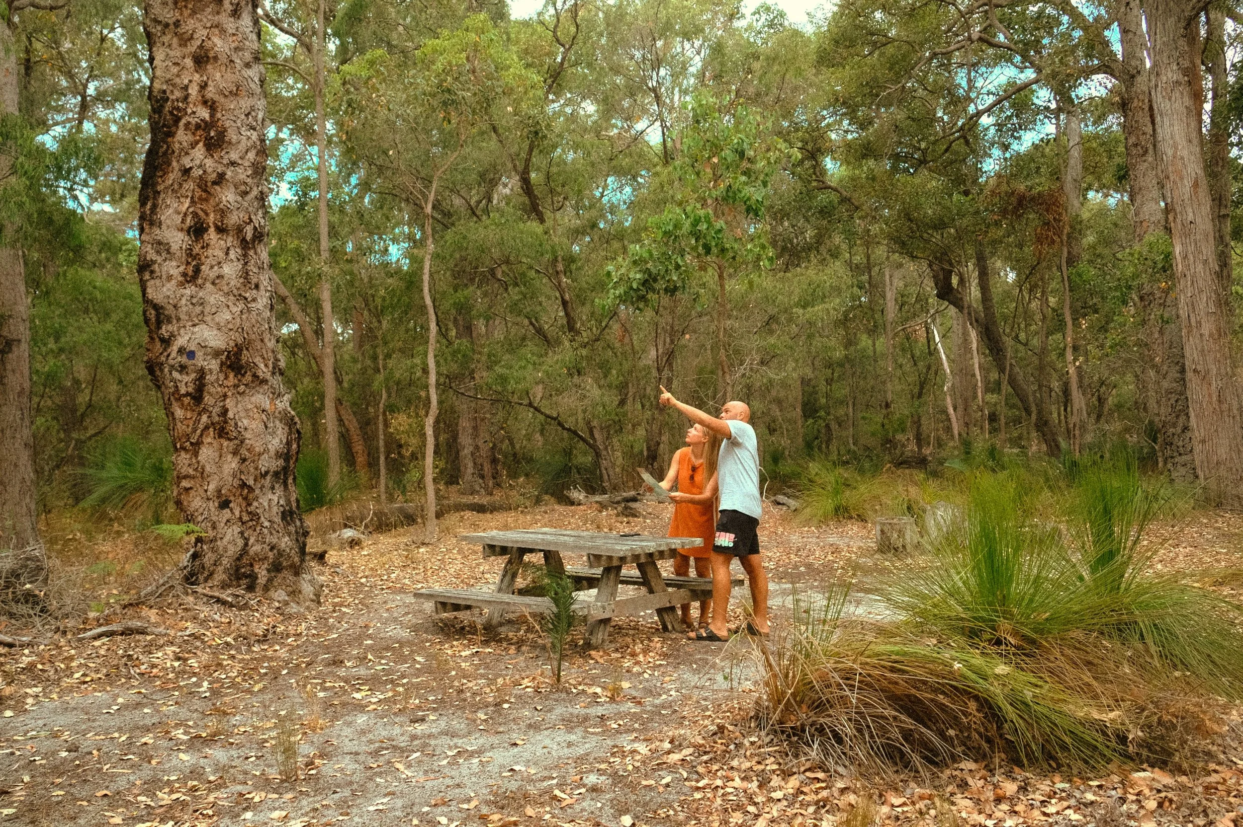 Two people standing next to a picnic table in a wooded outdoor area at Bussells Bushland Cottages in Margaret River., with trees and dry leaf-covered ground surrounding them. 