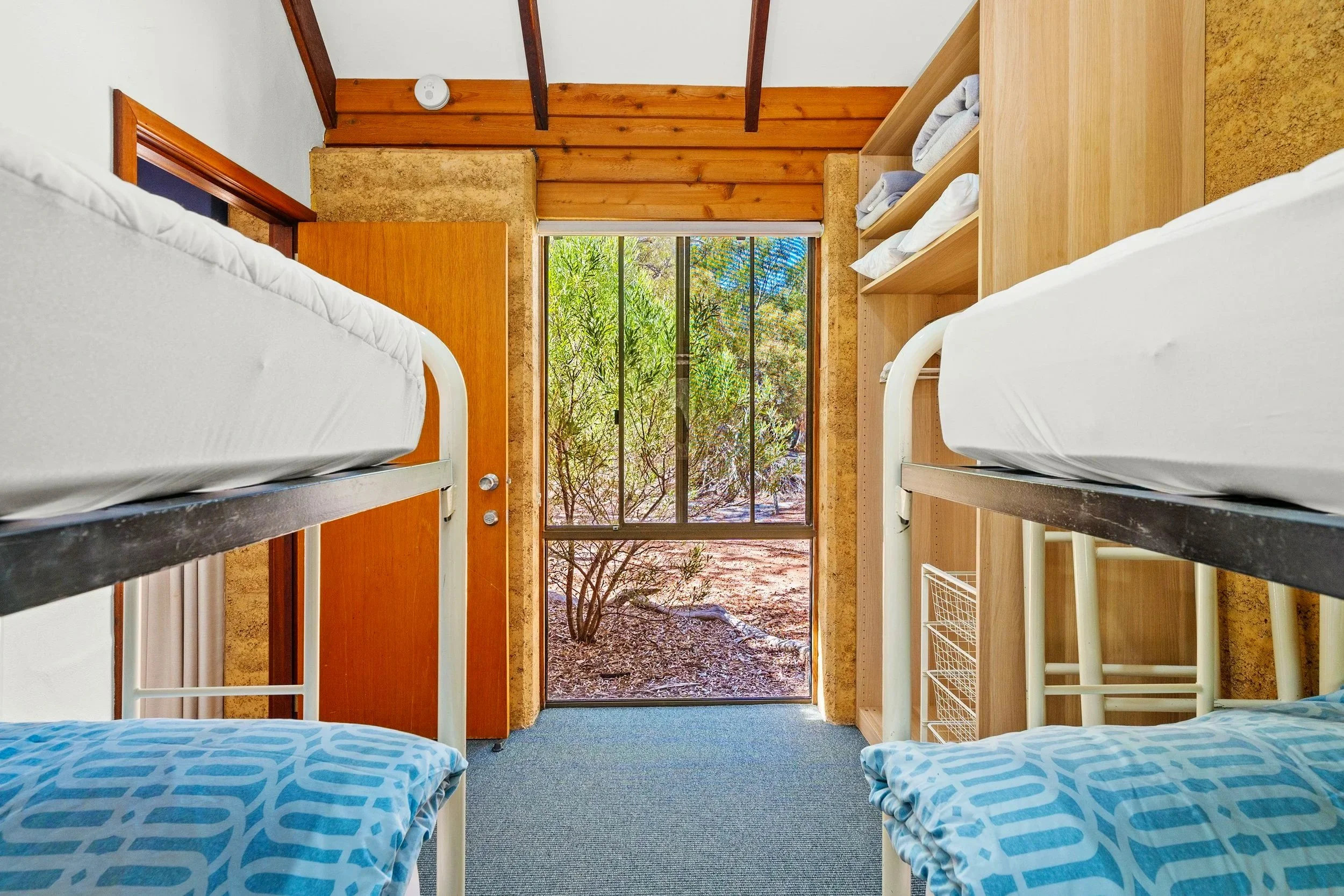 Room with four bunk beds, two on each side, with white linens and blue bed covers, wooden shelves with towels, a door opening to an outdoor garden with bushes and trees, and a window with metal bars.