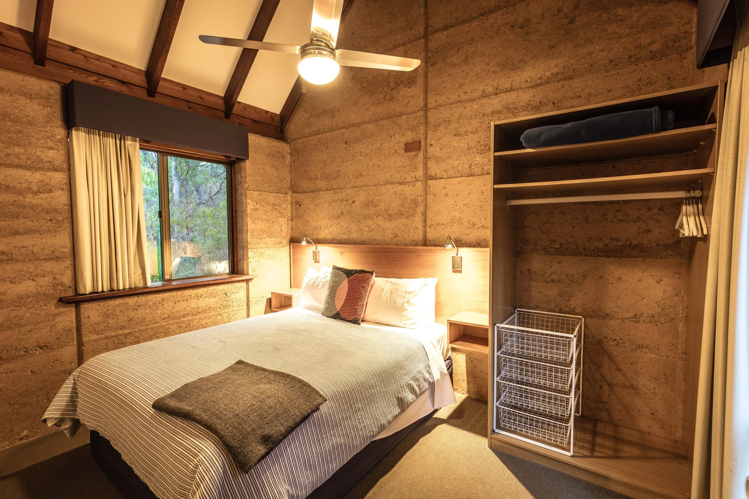 A cozy bedroom with a large window and a queen bed at Bussells Bushland Cottages in Margaret River Australia. There is a nightstand with reading lamps on either side of the bed, and a wooden open closet on the right with folded blankets and hangers.