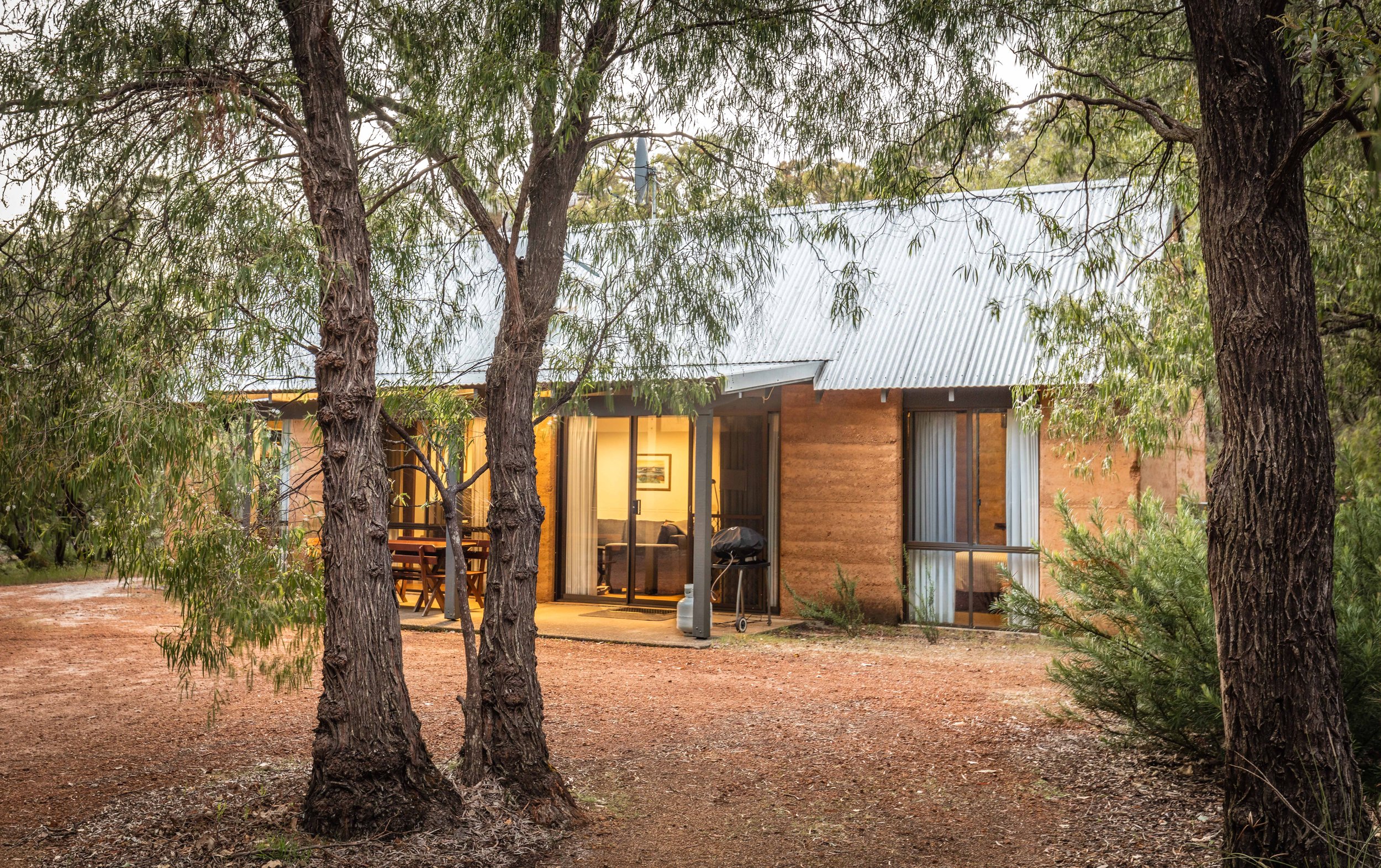 A cozy house at Bussells Bushland Cottages in Margaret River nestled among trees with outdoor patio and barbecue grill, illuminated from inside.