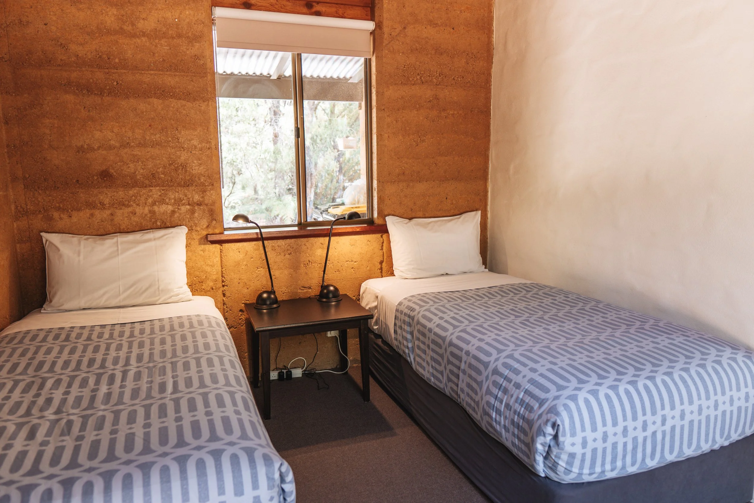 Bedroom with two beds, a window, and a small black nightstand with two black lamps, in a room with beige and brown walls.