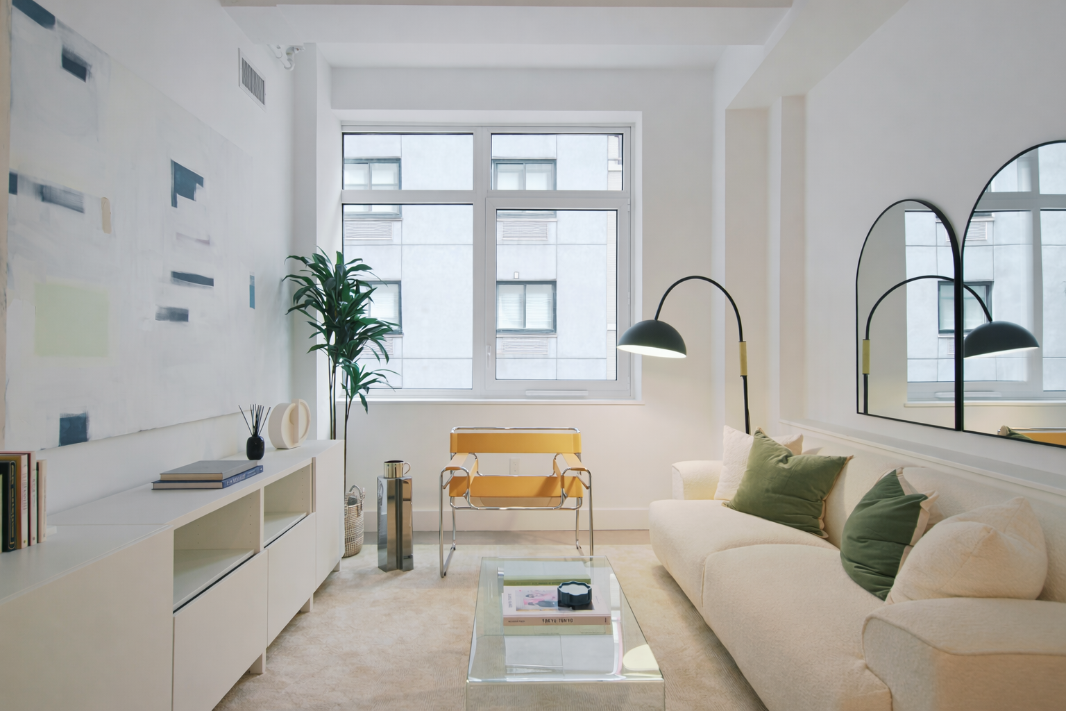 Bright living room with white walls, large window, white sofa with green cushions, glass coffee table, black arc floor lamps, large mirror, yellow chair, indoor plant, and colorful abstract art on wall.