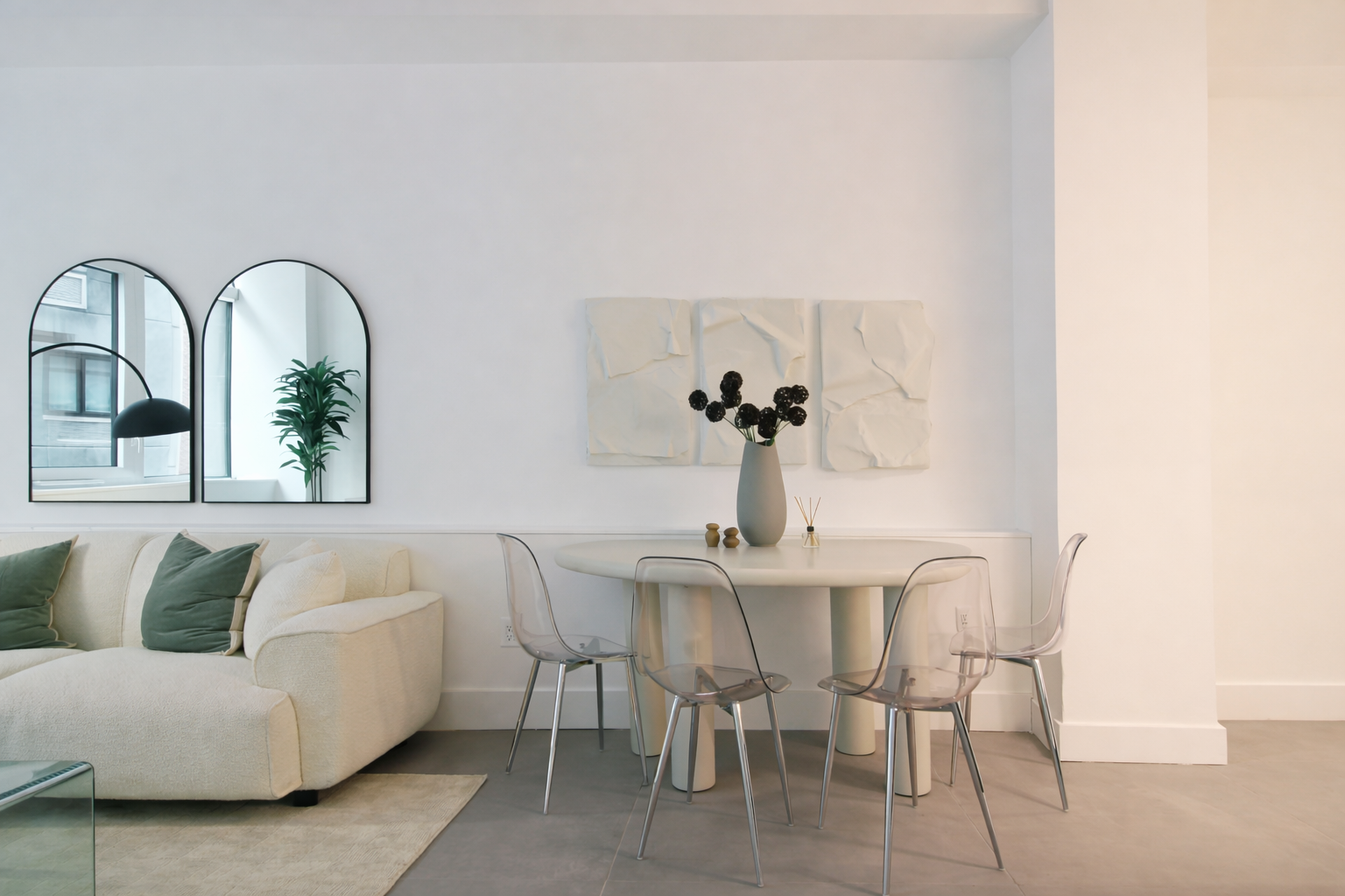 Minimalist living room with white couch, green and white pillows, three arched mirrors on wall, white dining table with four translucent chairs, black vase with black flowers, and white abstract wall art.