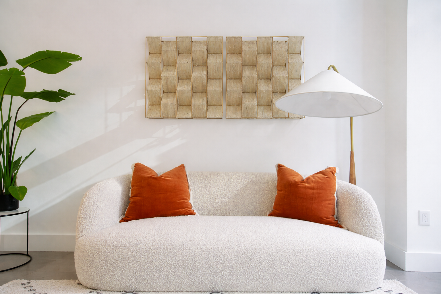 Living room with white textured sofa, two orange pillows, a large floor lamp with a white shade, a large potted plant on a small black table, and decorative wall art on a white wall.