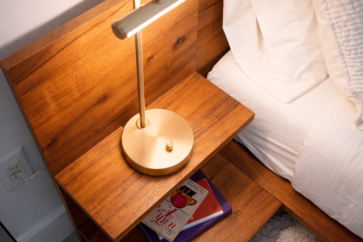 Wooden nightstand with a brass-colored table lamp and a bed with white bedding.