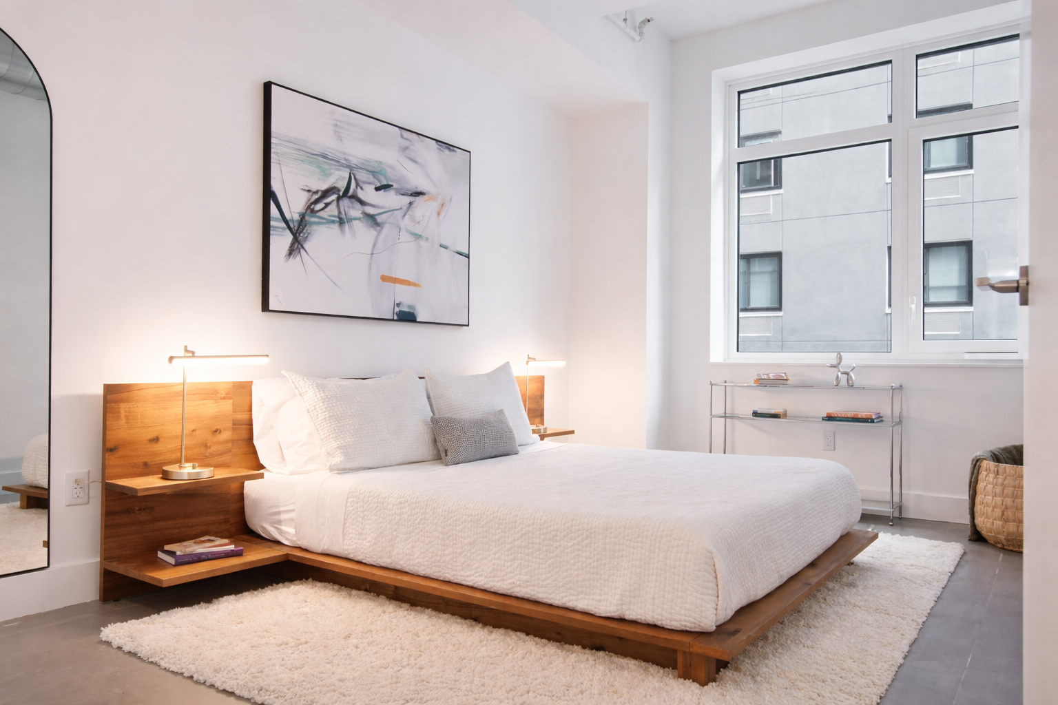 Modern bedroom with a wooden bed frame, white bedding, and a large abstract painting above the bed. There are two small nightstands with lamps, a plush white rug, and a metal shelf near the window holding books. The room has large windows letting in natural light.