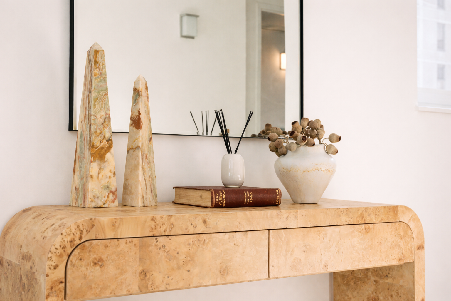 Decorative wooden console table with two tall marble sculptures, a white vase with black reed diffuser sticks, an open vintage book, and a white vase with dried plant stems, mirrored wall and window in background.
