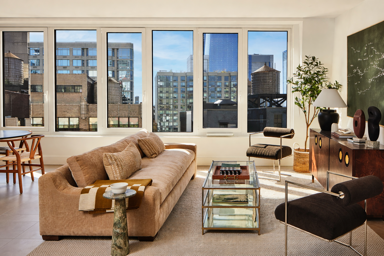 Living room with large windows showing city buildings, beige sofa with cushions, glass coffee table, black and brown chairs, and a sideboard with decorative objects and a lamp.