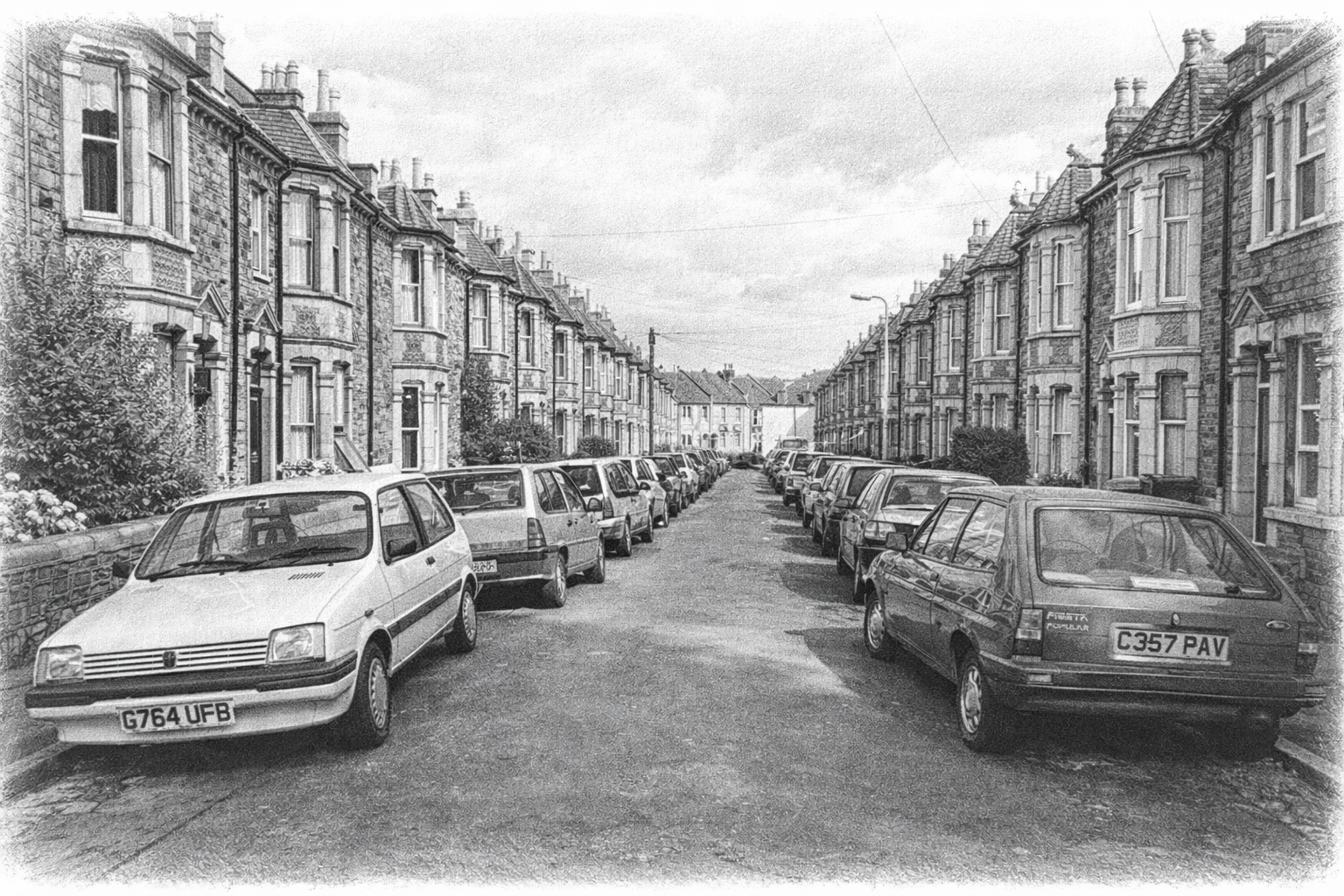 A black-and-white photograph of a residential street lined with old stone houses and numerous parked cars on both sides of the street.