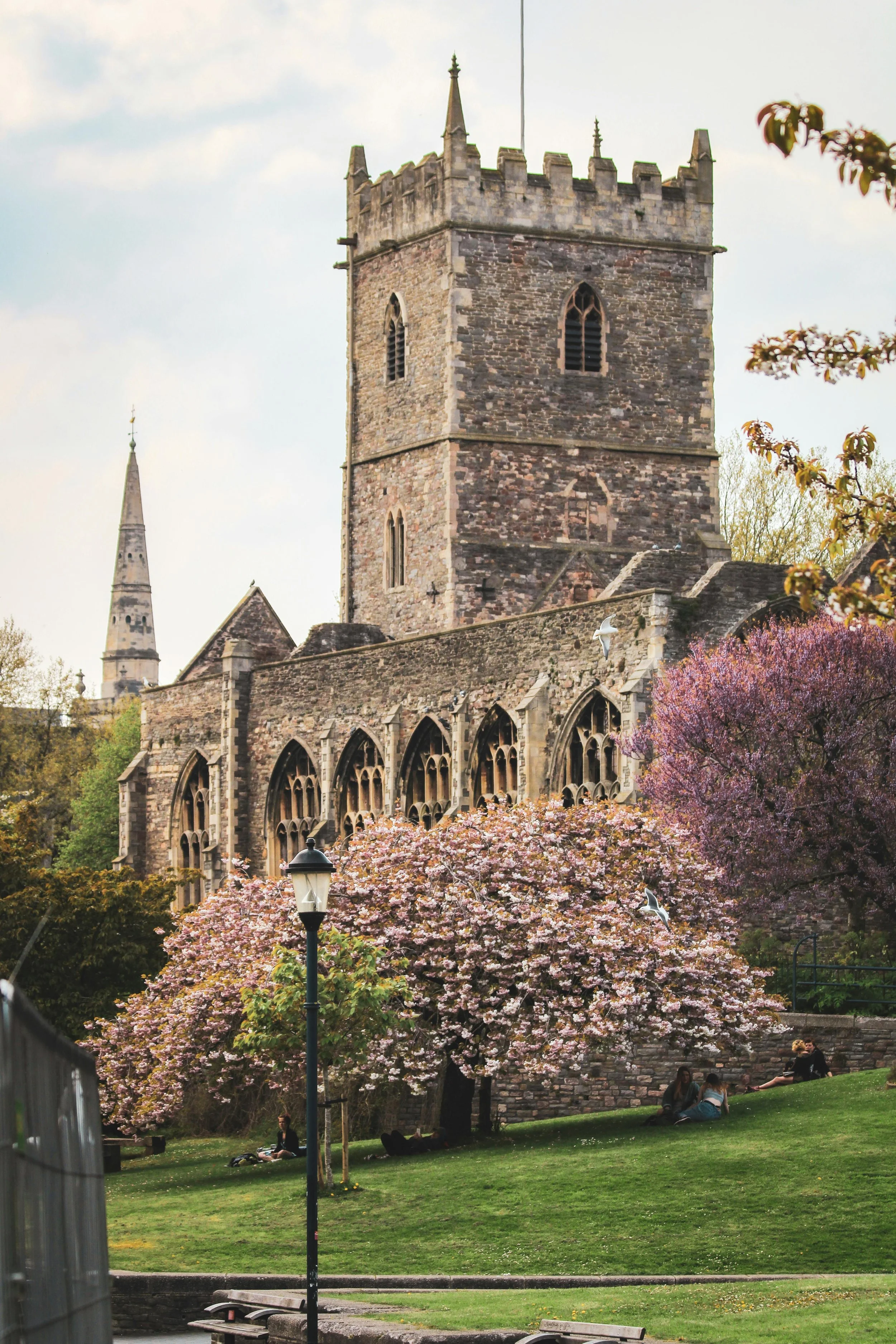 A historic stone church with a tall square tower, gothic windows, and battlements, surrounded by blooming pink and purple trees, with a few people sitting on the grass and walking nearby.