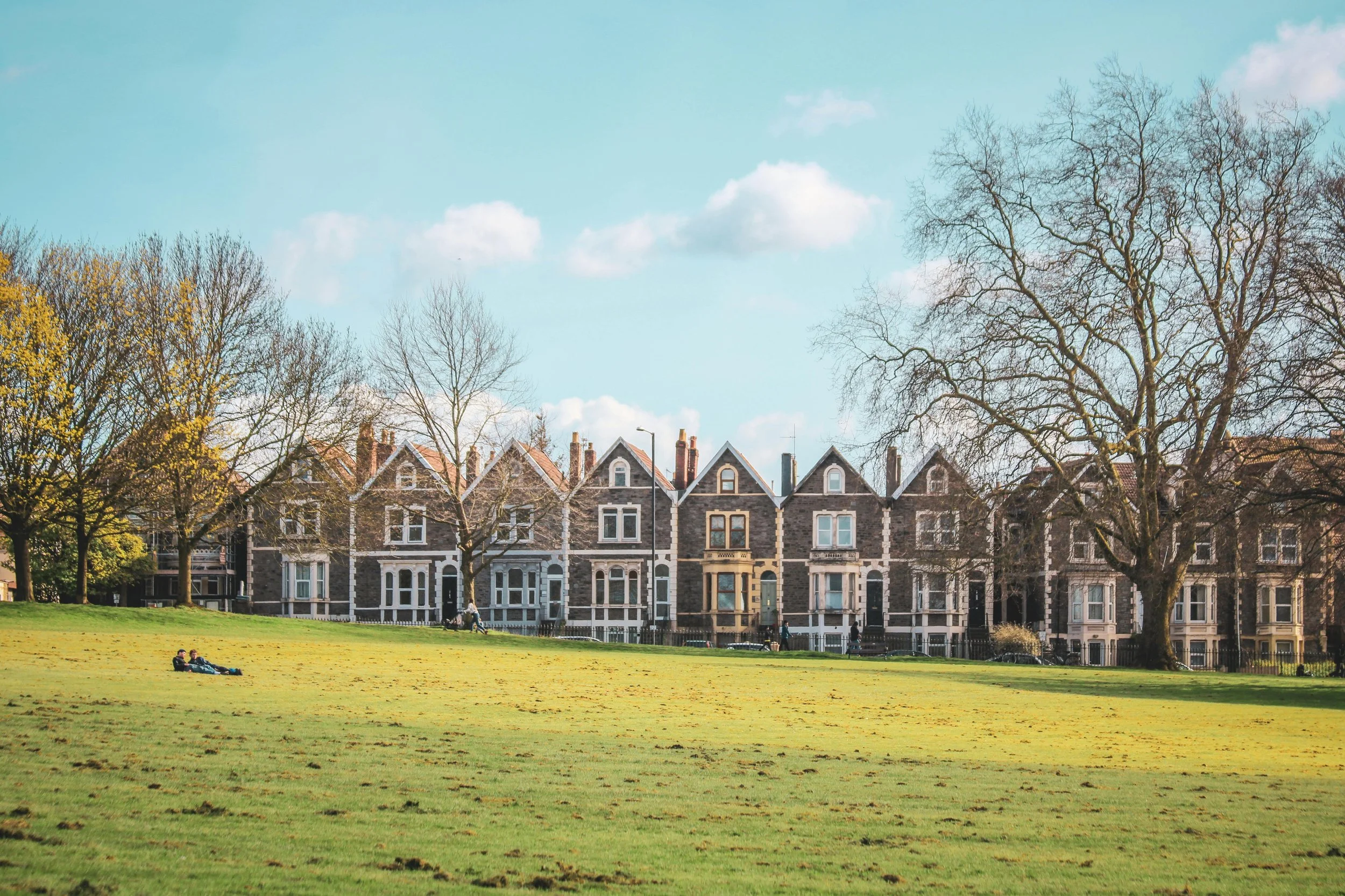 A row of gray stone Victorian houses with pointed roofs behind a grassy park. Several leafless trees are in the park, and a couple of people are sitting on the grass.