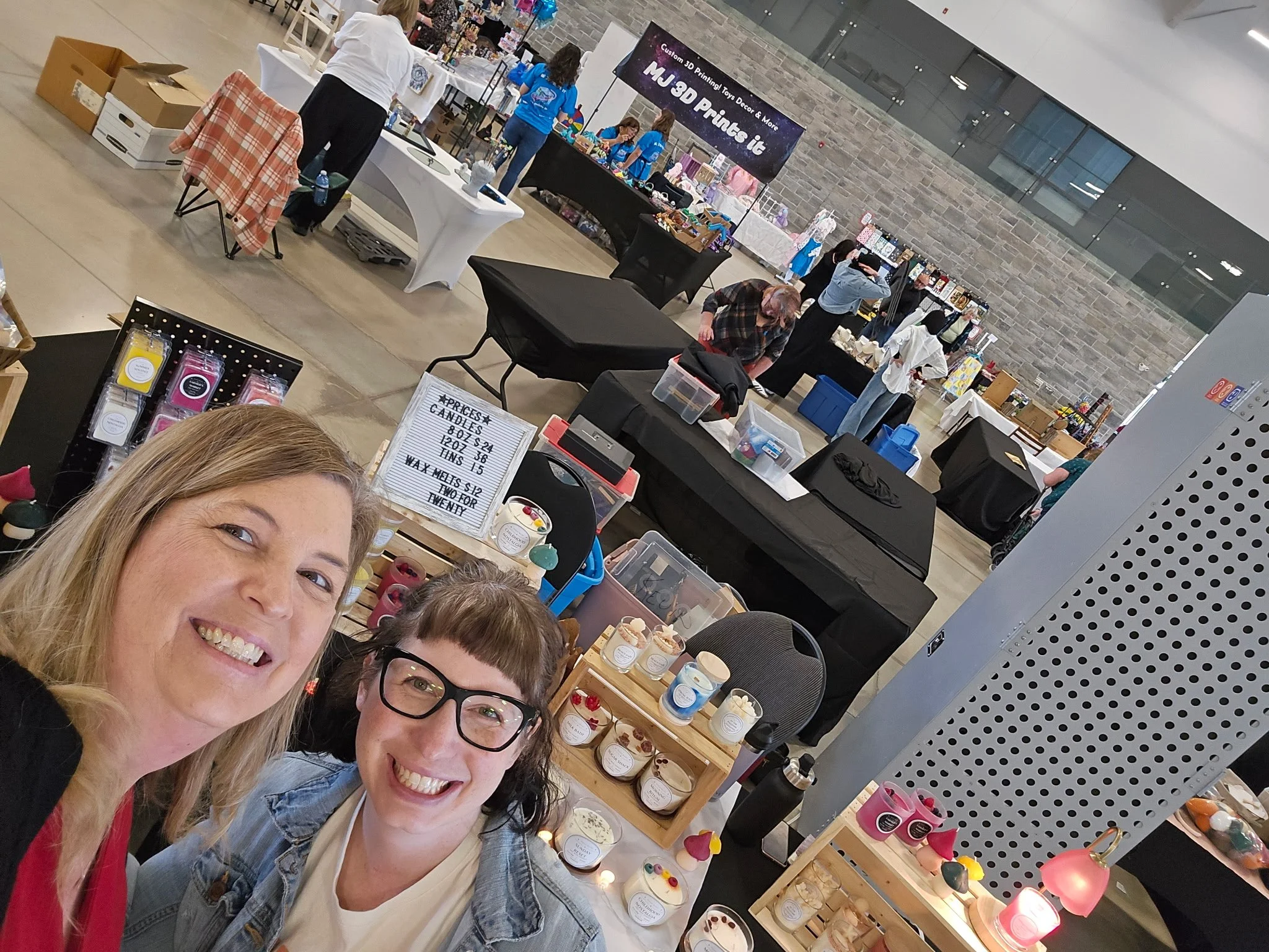 People taking a selfie at an indoor craft fair or market with booths displaying various handmade items in the background.