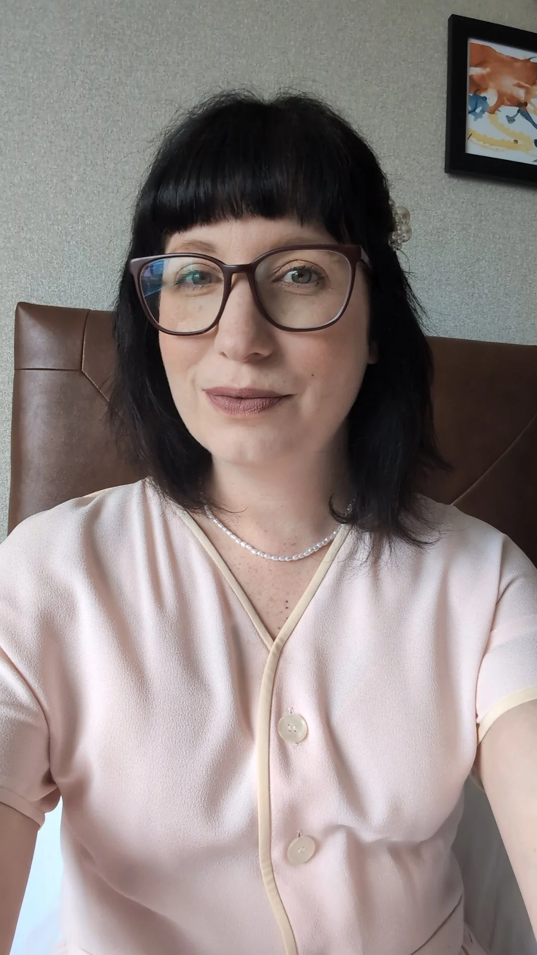 A woman with black hair and glasses sitting in front of a beige wall, wearing a light pink top and a pearl necklace.