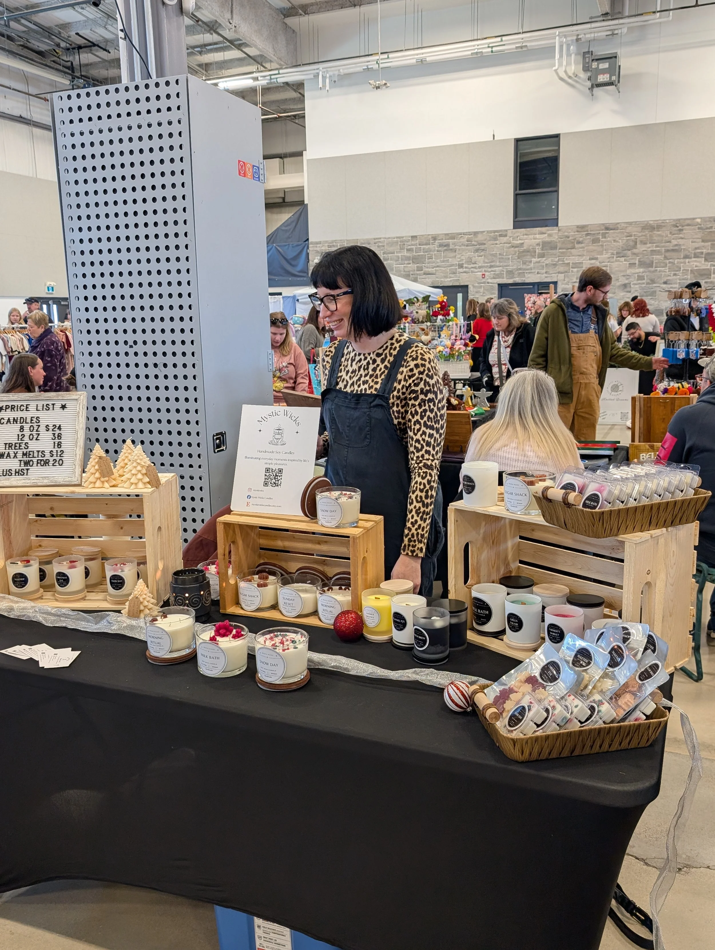 A woman with black hair and glasses wearing a leopard print shirt and apron standing behind a table displaying various candles in a busy indoor market or craft fair. The table has a black cloth and wooden display boxes with candles and small decorative items, with other shoppers and booths visible in the background.