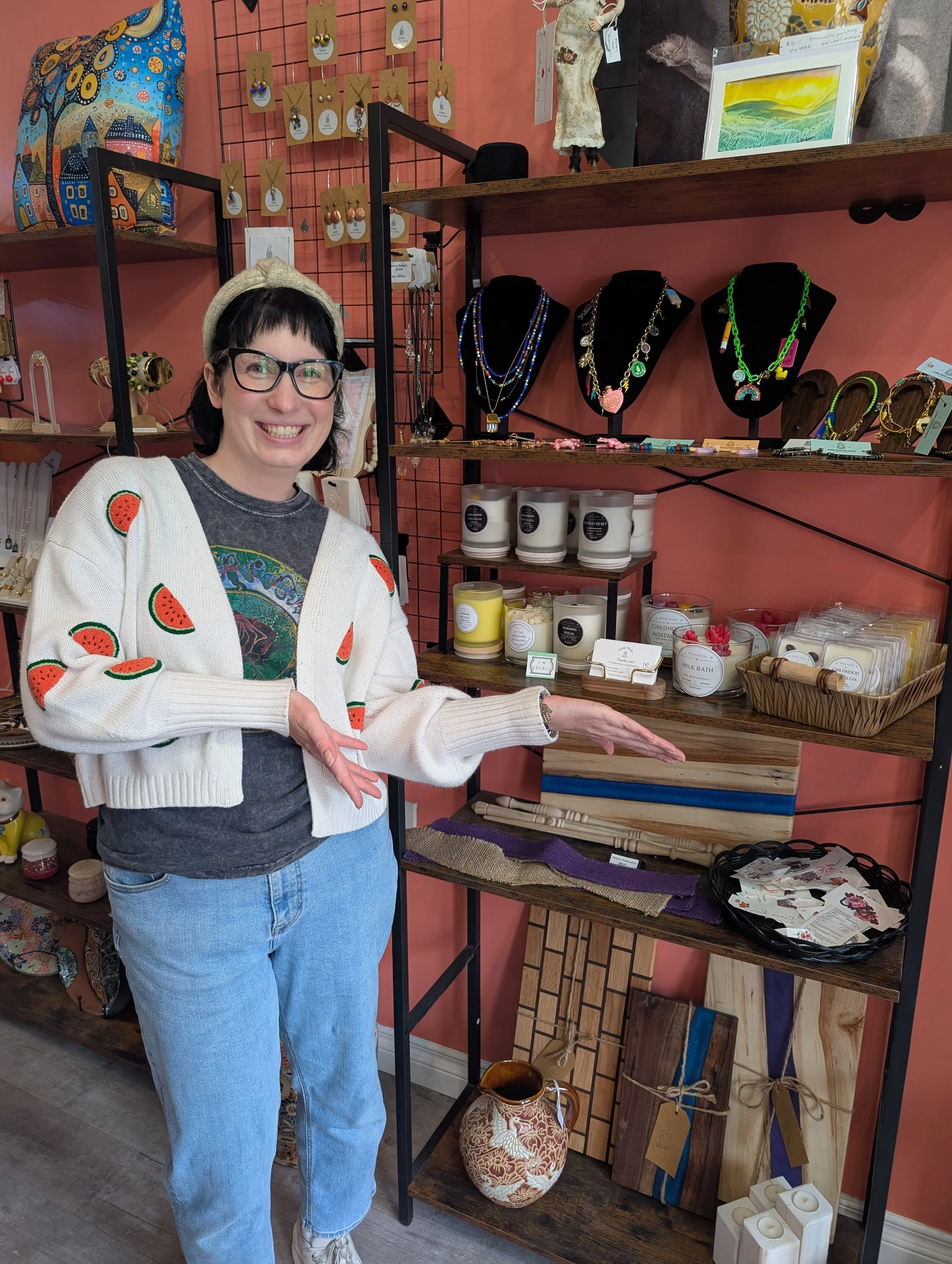 A woman with short black hair, glasses, and a beige headband is smiling and pointing to a display of jewelry and crafts in a store with a pink wall. She is wearing a white cardigan with watermelon embroidery over a gray T-shirt and light blue jeans.