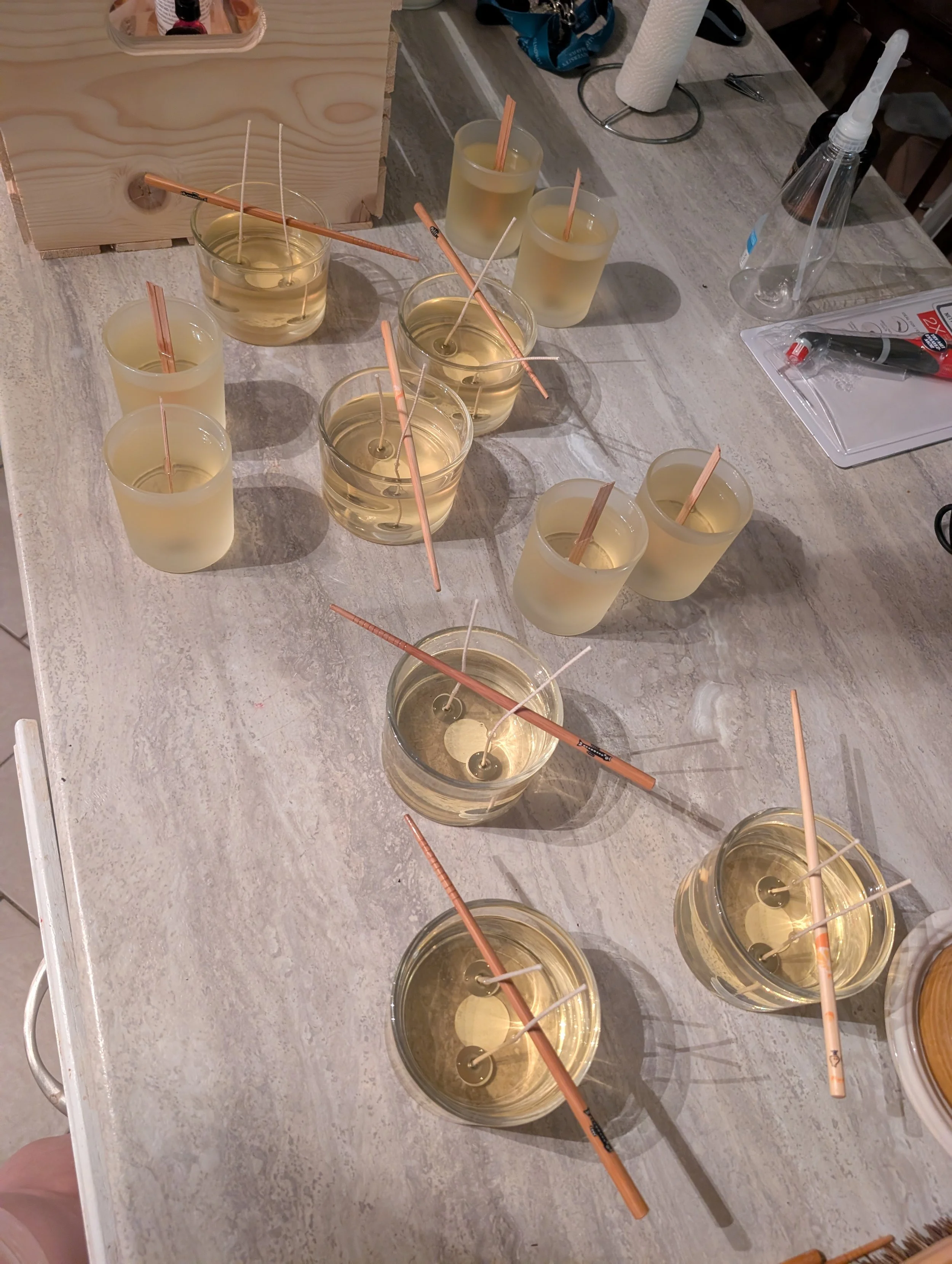 Several glasses filled with liquid wax, each with a candle wick, Scattered around on a light-colored wooden table are additional wicks, a tissue holder, a paper towel roll, a plastic bottle, and some tools or utensils.