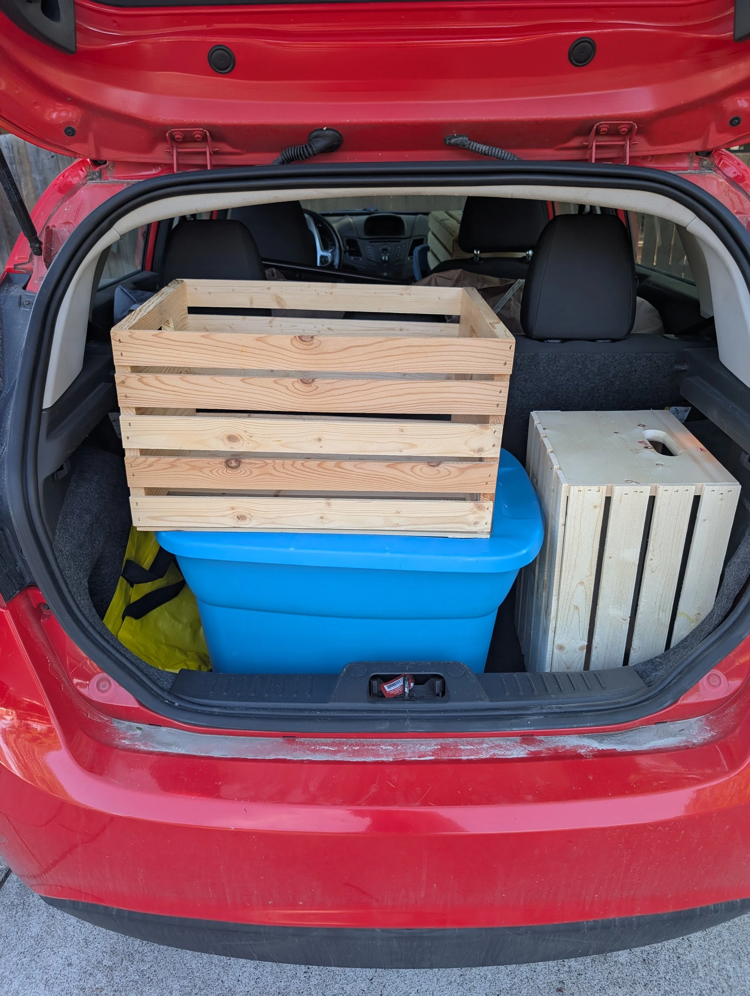 The open trunk of a red car showing a large blue plastic storage bin, two wooden crates, and other miscellaneous items inside.