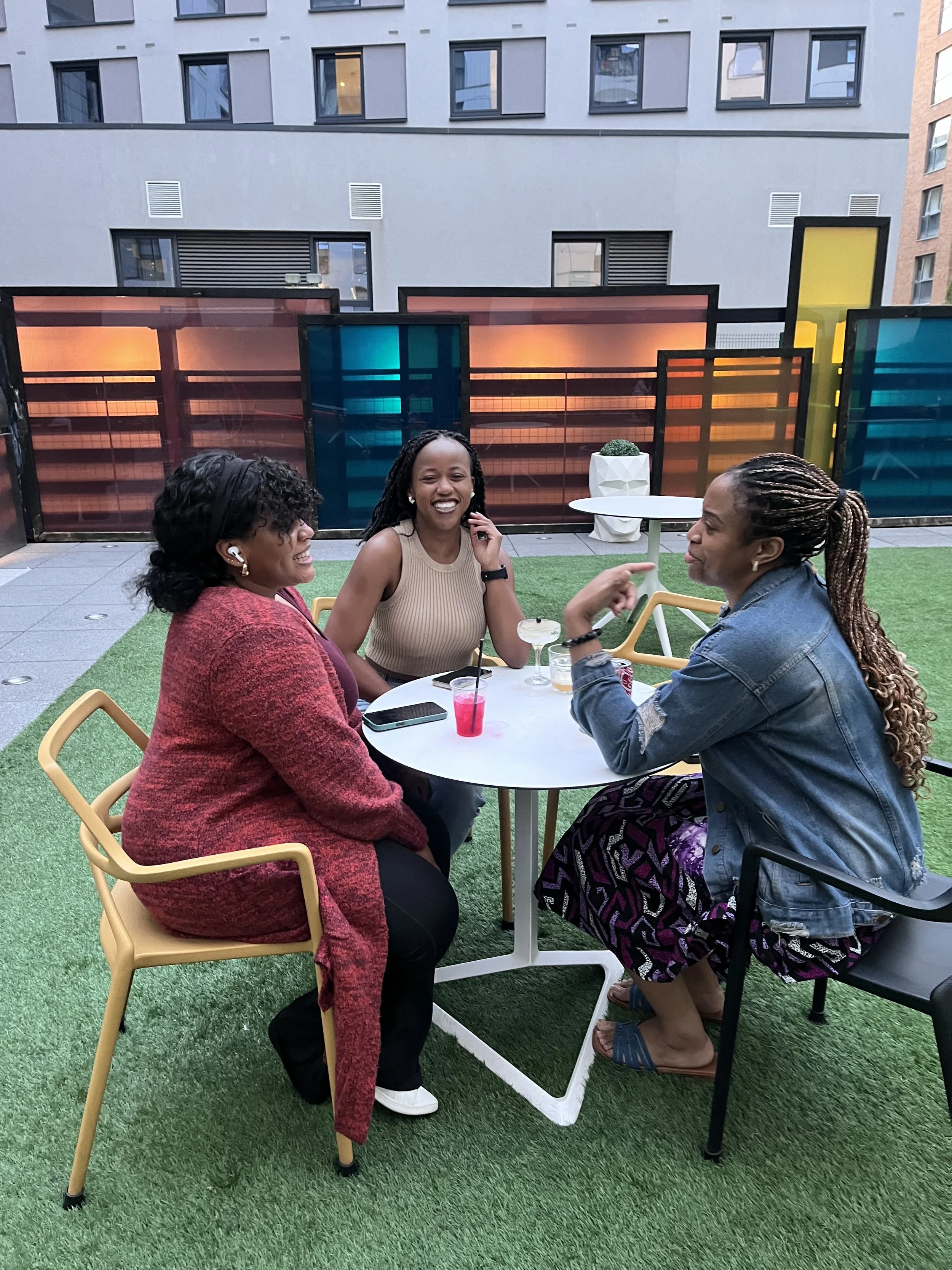 Three women sitting at a round white table on a rooftop, enjoying drinks and conversation, with colorful translucent divider panels and a modern cityscape background.