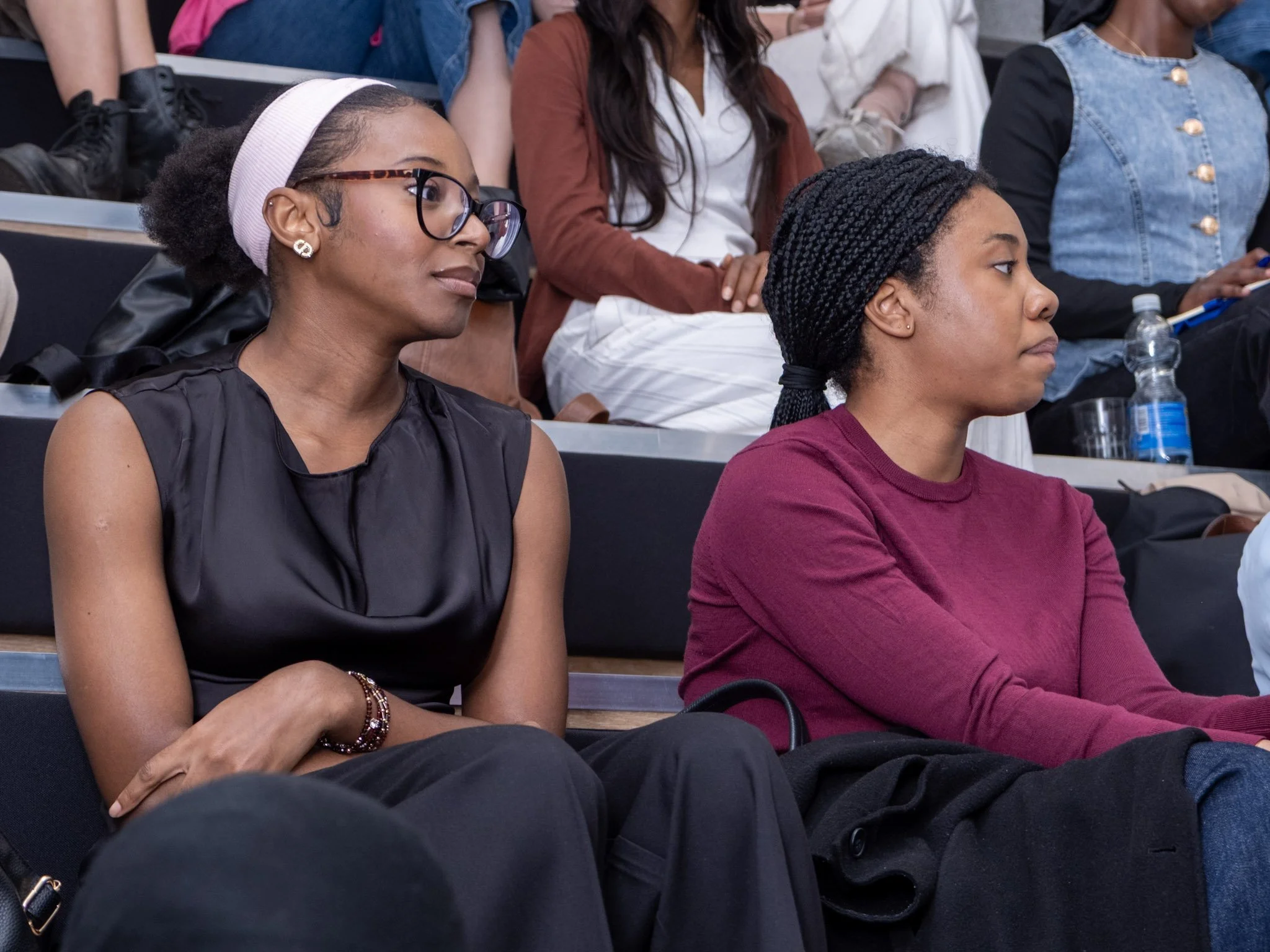 Two women sitting in a row of seats at an event, paying attention. The woman on the left has glasses, a pink headband, earrings, and is wearing a sleeveless black top. The woman on the right has braided hair, a maroon long-sleeve shirt, and appears focused on the event.