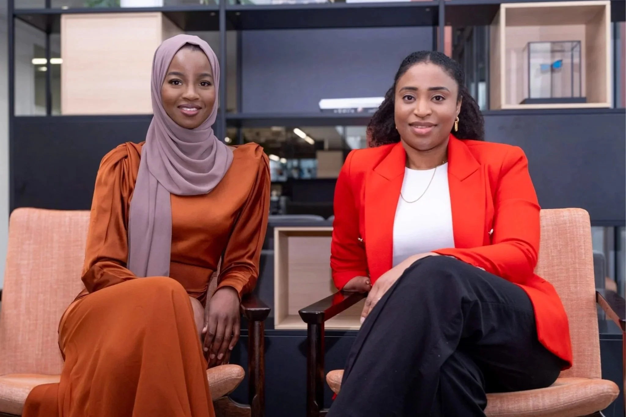 Two women sitting on chairs in a modern office space, smiling at the camera. One woman is wearing a hijab and a rust-colored dress, while the other is dressed in a red blazer and black pants.