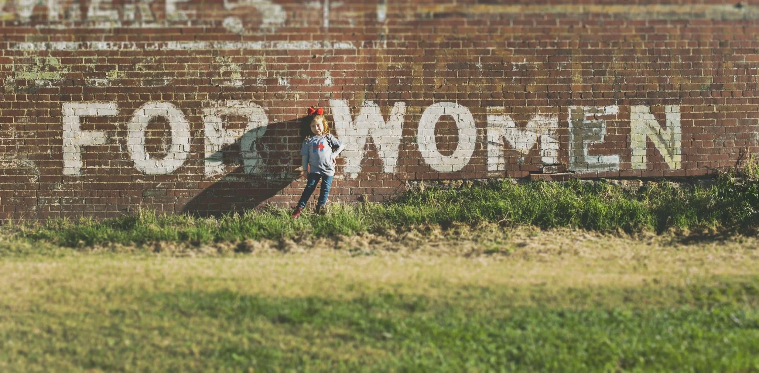 A young girl with red hair, wearing a grey jacket and blue jeans, stands in front of a brick wall with the word 'FOOTMONEN' painted in white. She is next to her shadow, which is cast on the wall. The grass in front of the wall is green and slightly overgrown.