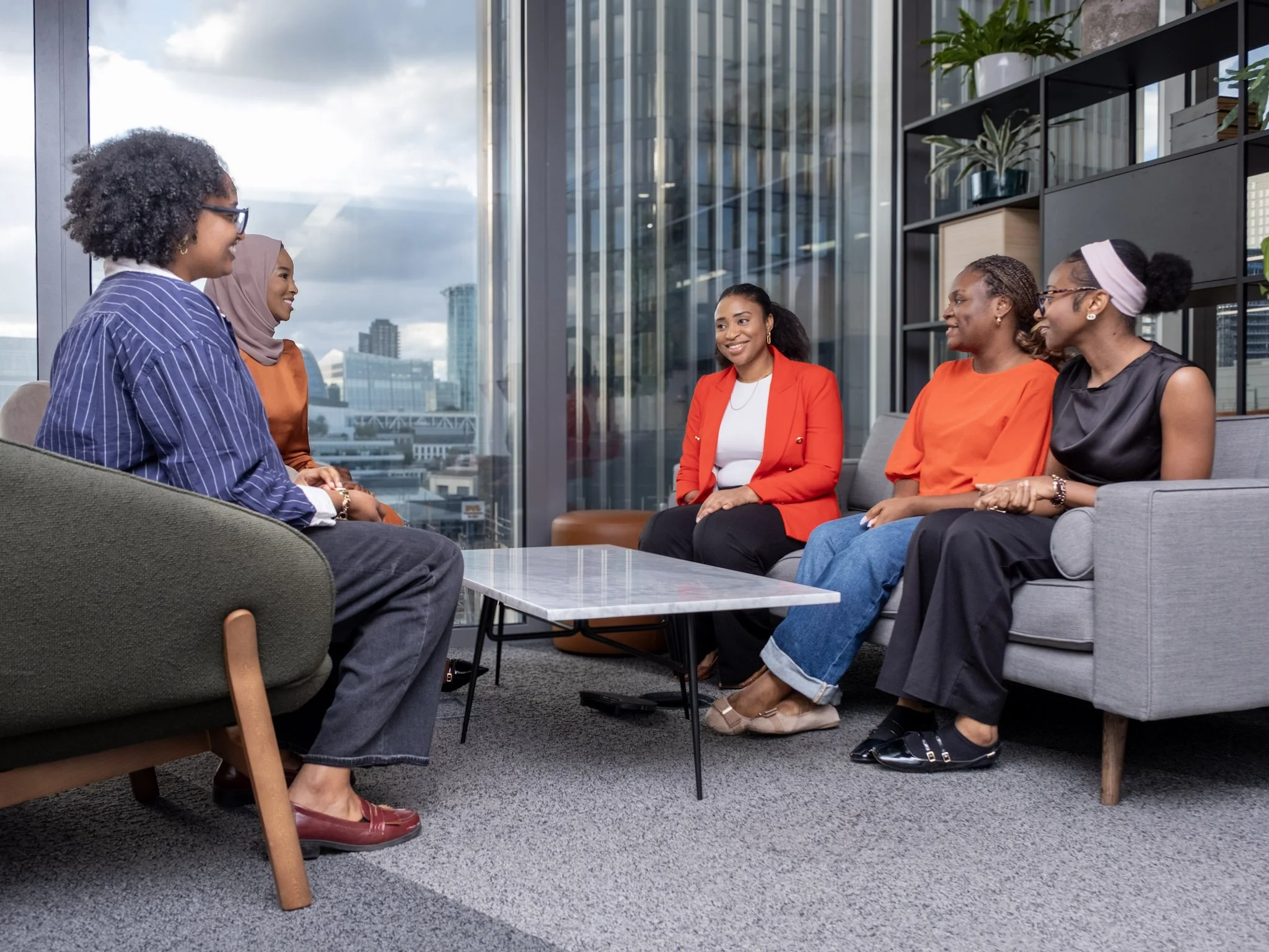 Five women sitting and talking in a modern office lounge with large windows and city view.