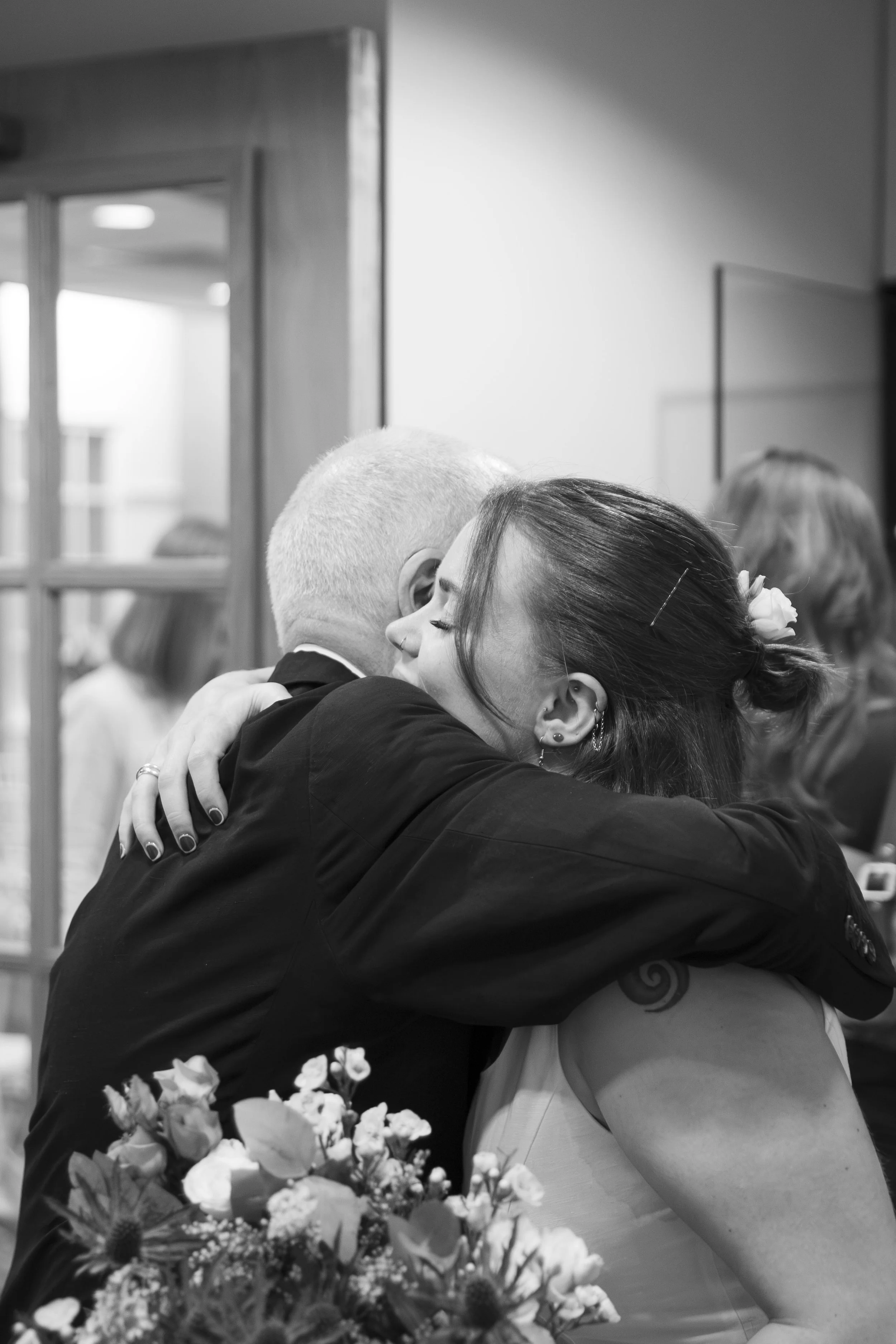 A couple sharing a kiss at a wedding, with flowers in the foreground and people in the background.