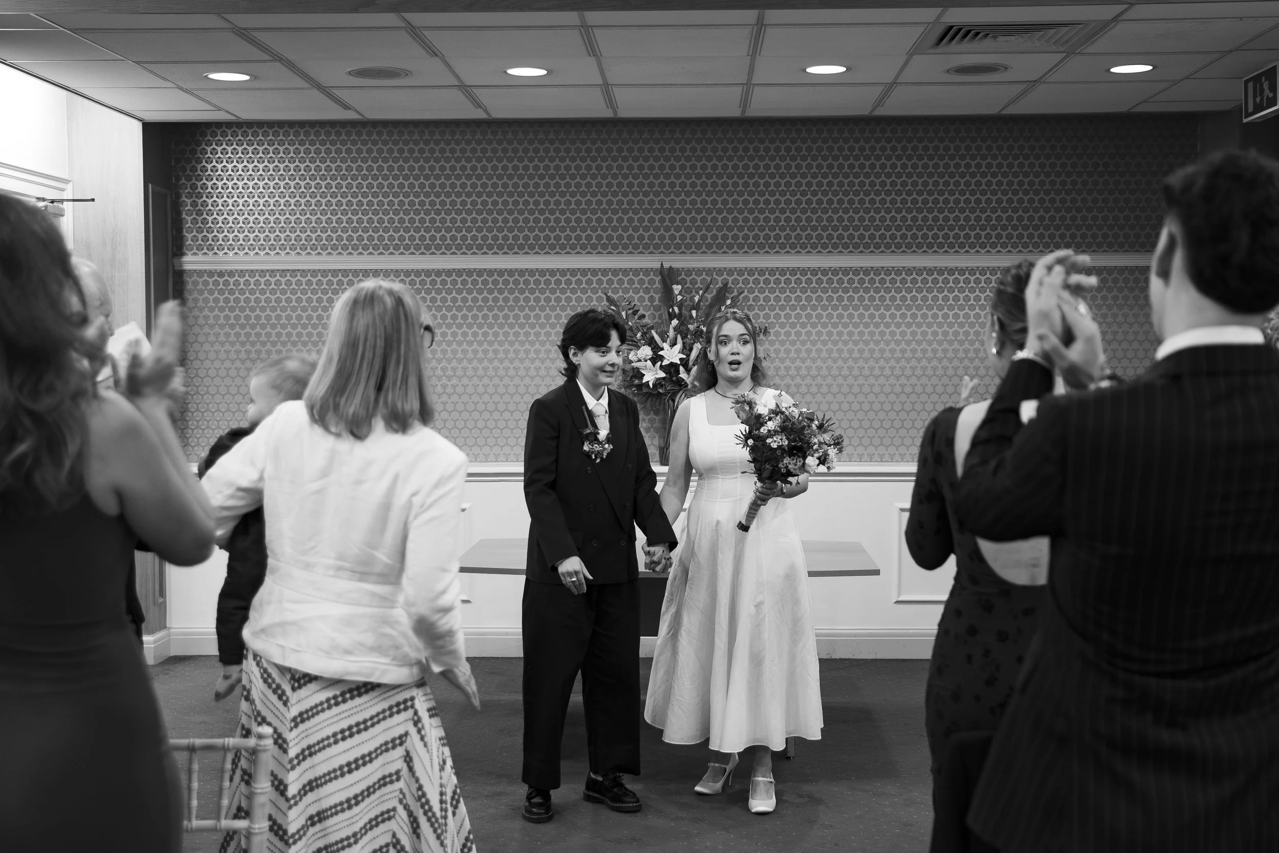 A bride and groom at their wedding ceremony, holding hands while holding a bouquet of flowers, with guests surrounding them inside a decorated room.