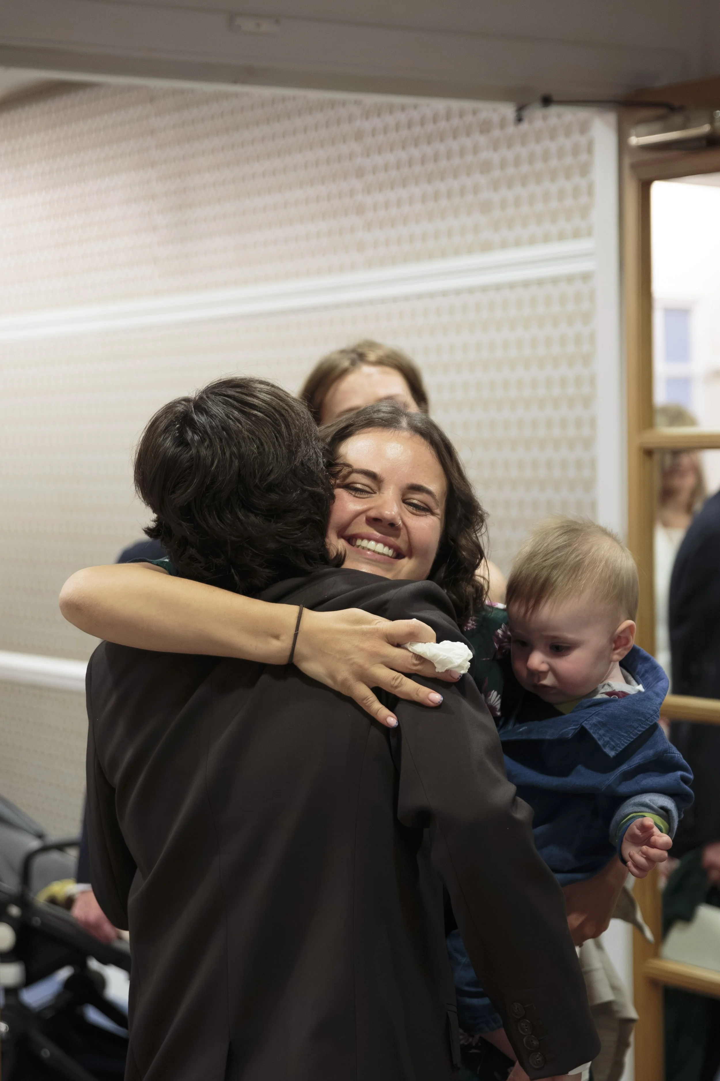 Several people hugging, with a woman smiling broadly in the center. Some children are present, one being held by the woman, in an indoor setting with a patterned wall and window in the background.