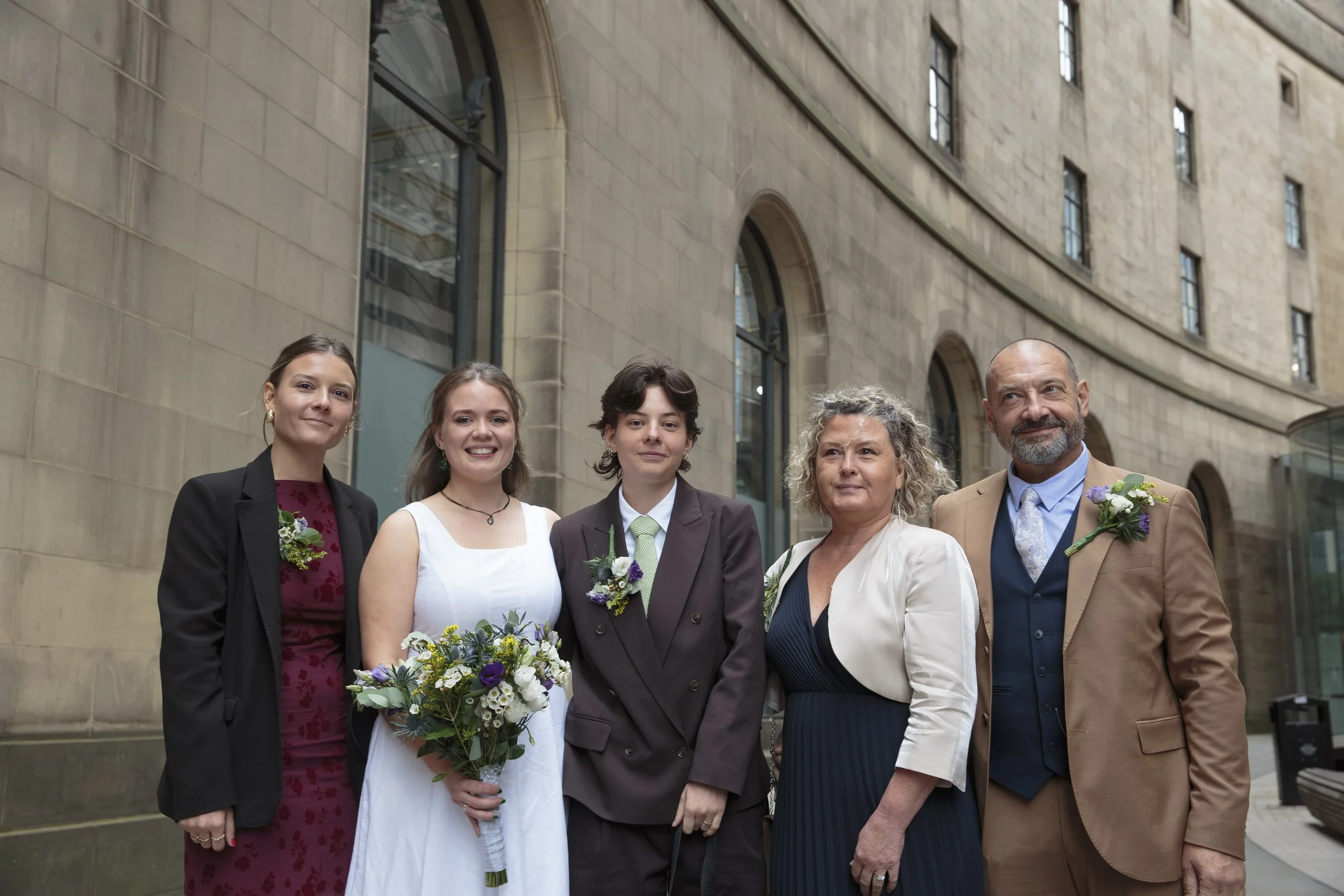 Group of five people standing outdoors in front of a stone building, dressed in formal attire, with one woman holding a bouquet of flowers.