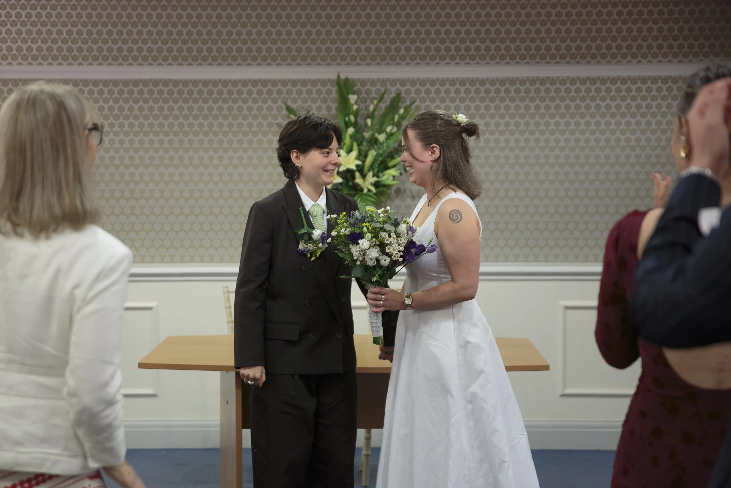 Two women at a wedding, holding hands and smiling at each other, with large floral arrangements and guests in the background.