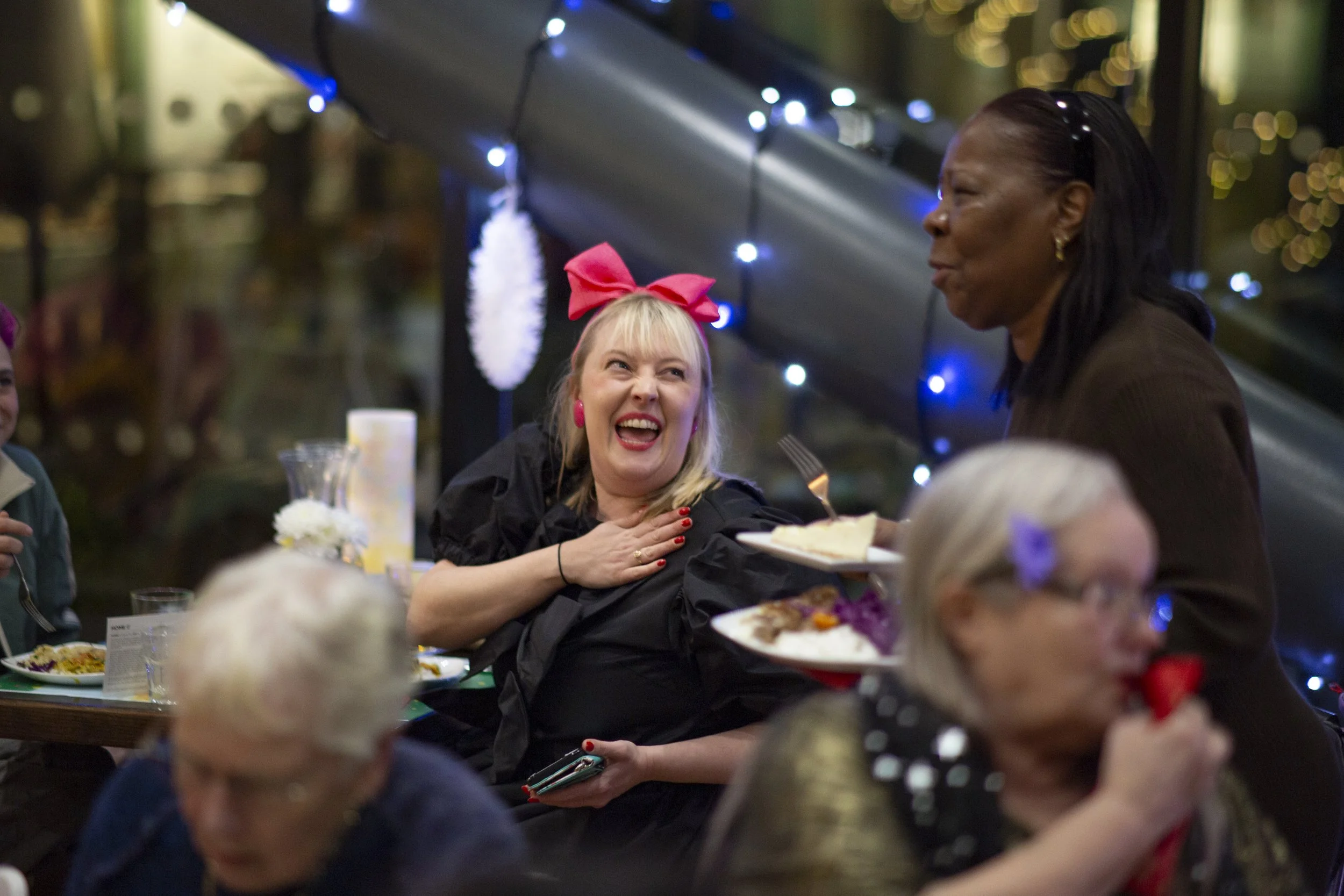 A woman with a pink bow in her hair laughing while sitting at a dinner table with others, a server is bringing her a plate of food, decorated with festive holiday lights in the background.