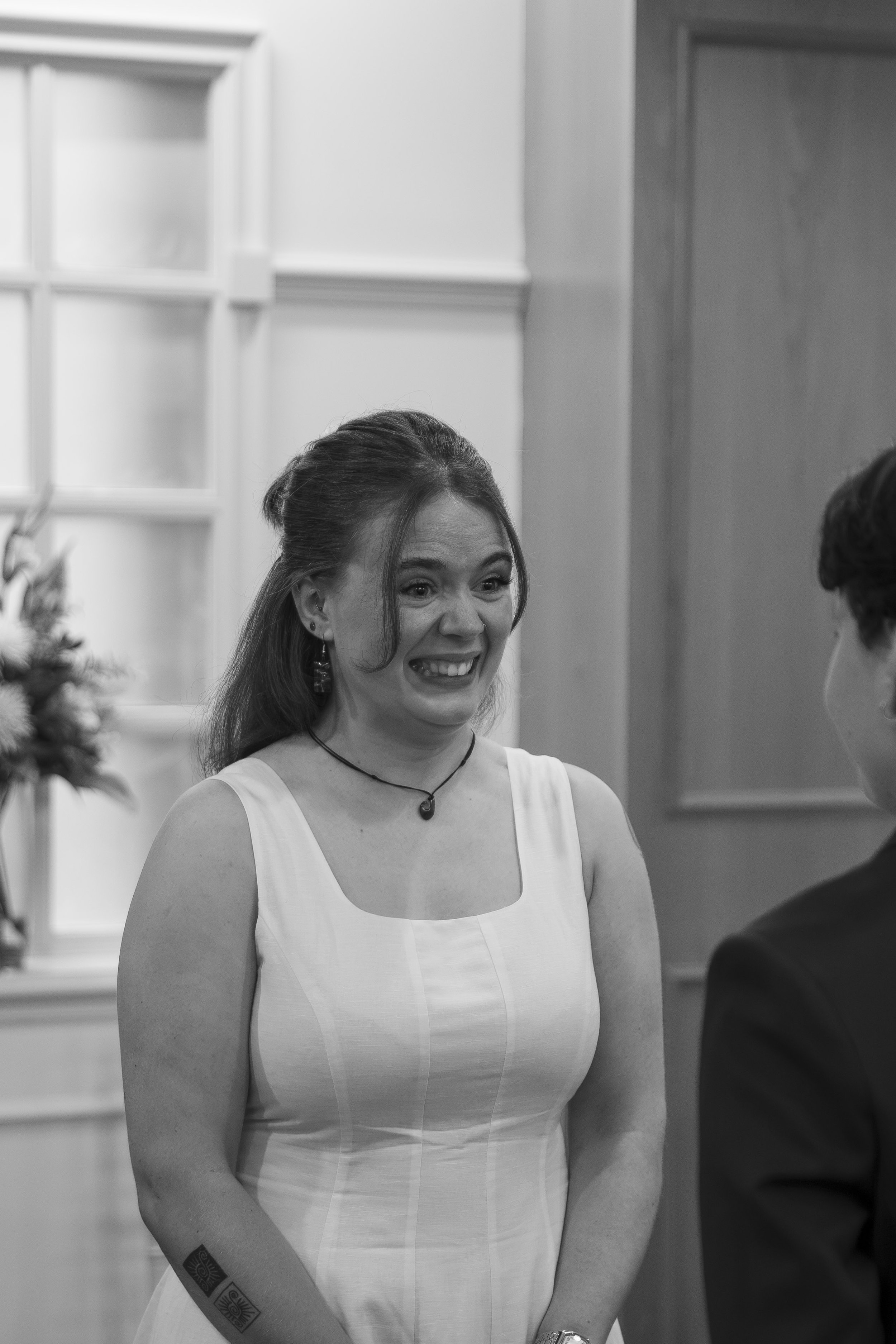 A woman with dark hair and earrings smiling, wearing a sleeveless white dress, interacting with a man in a suit in an indoor setting.