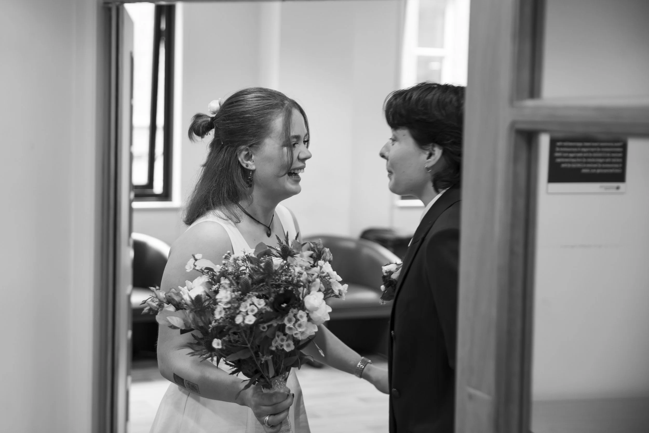 A woman in a wedding dress holding a bouquet, smiling, and looking into the eyes of a man in a suit, in a room with windows, seen through a partially open door.