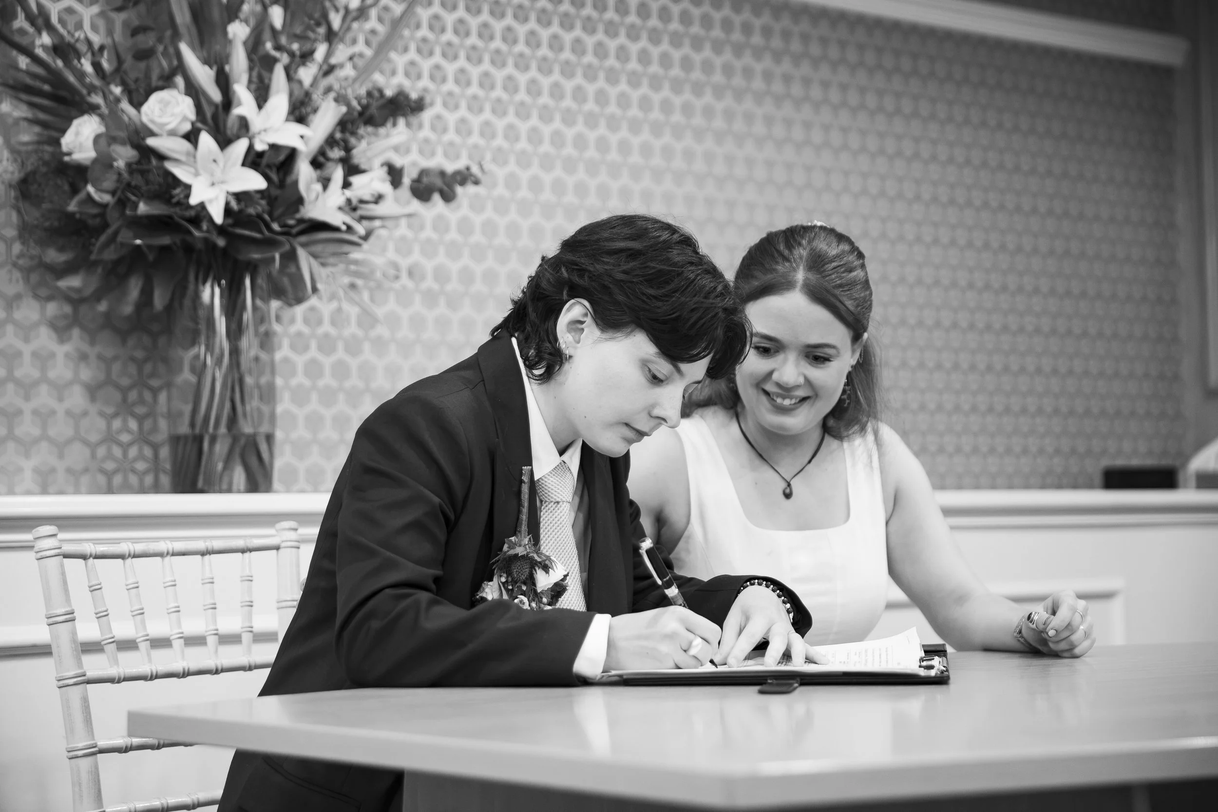 A man and a woman sitting at a table, signing a document, with a large bouquet of flowers in the background.