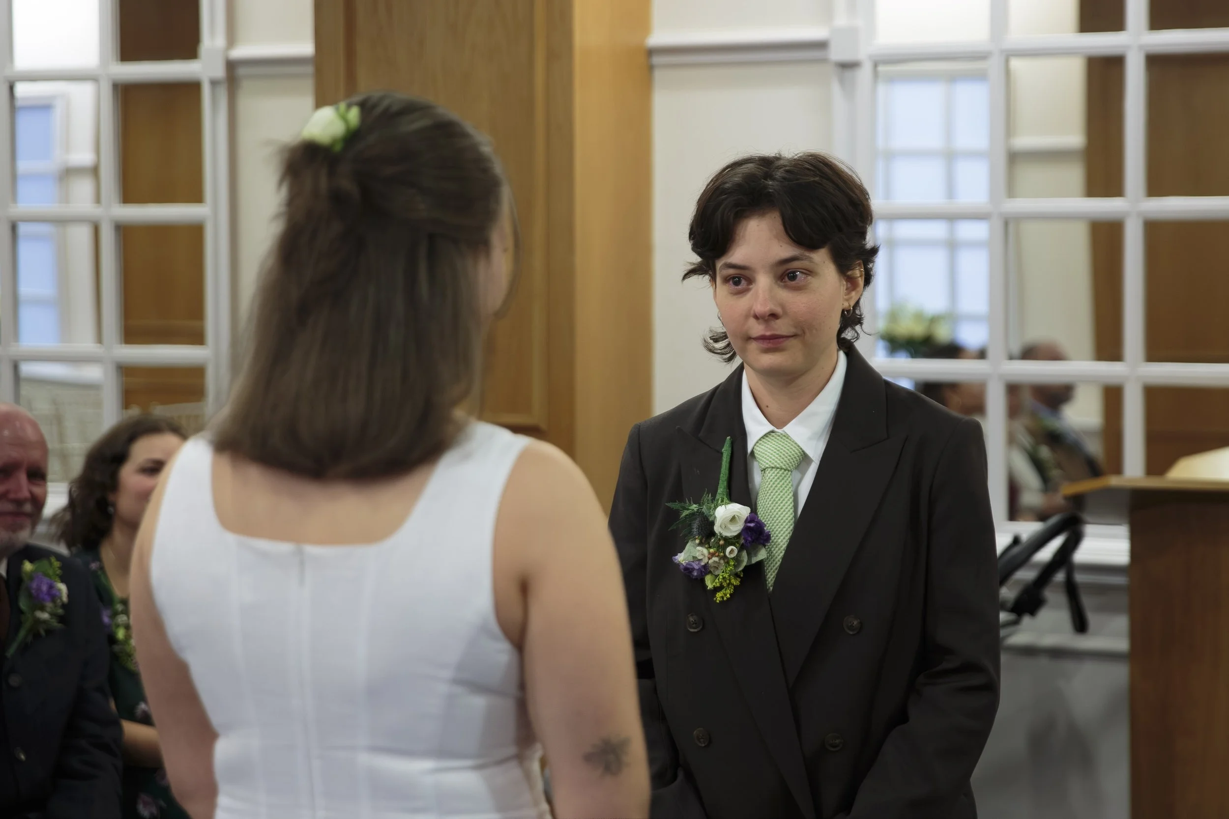A woman with short dark hair and a boutonniere on her jacket looks at another woman with long brown hair wearing a white dress, at a wedding ceremony in a bright indoor setting with wooden accents and large windows.