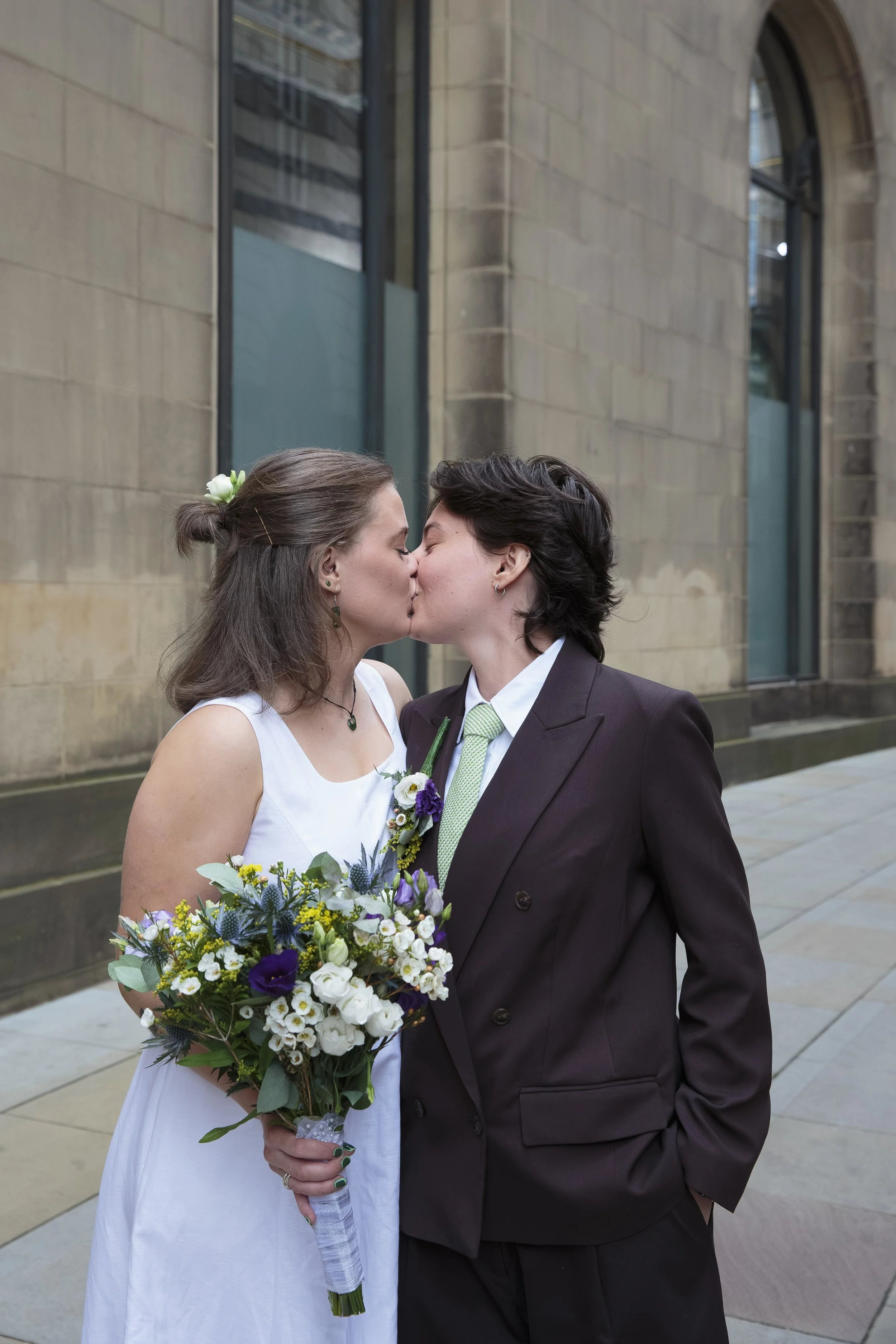 A couple in wedding attire sharing a kiss outdoors, with the bride holding a bouquet of flowers and wearing a white dress, and the groom wearing a dark suit.
